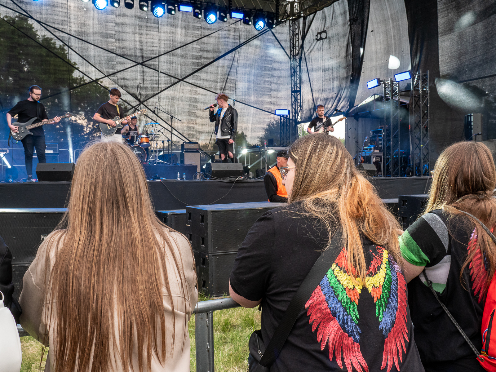 Na scenie odbywa się koncert - czterech wykonawców, mężczyzna śpiewa do mikrofonu, trzech gra na gitarach. Przed sceną przy barierkach trzy studentki.