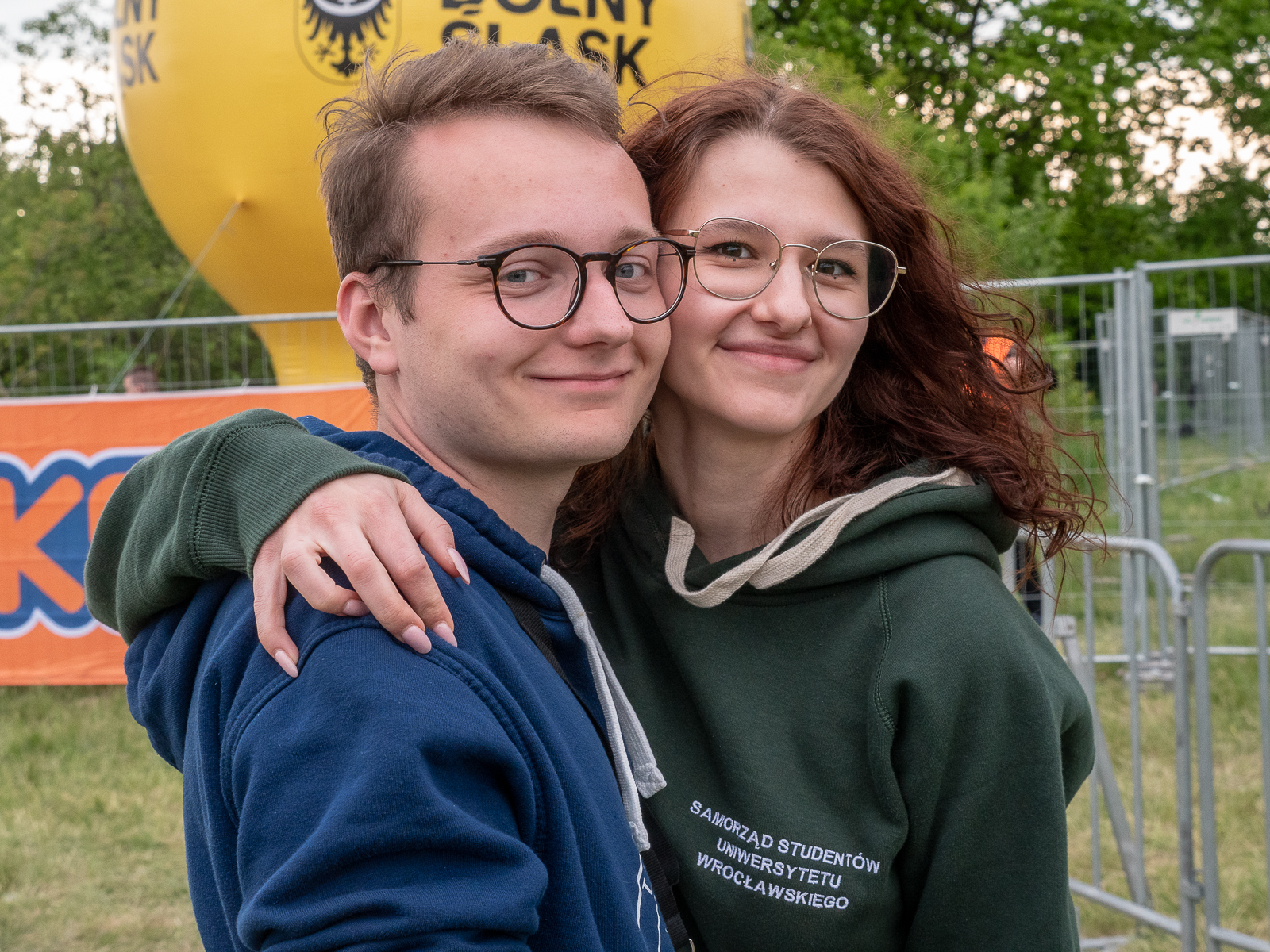 Student i studentka, uśmiechnięci, w okularach. Ona trzyma go za ramię, zbliżają się policzkami.