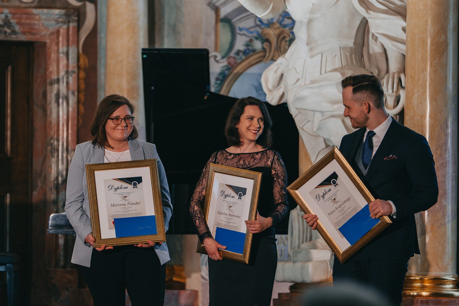 dwie młode kobiety i jeden mężczyzna w oficjalnych ubiorach stoją trzymając w rękach oprawione w złote ramy dyplomy