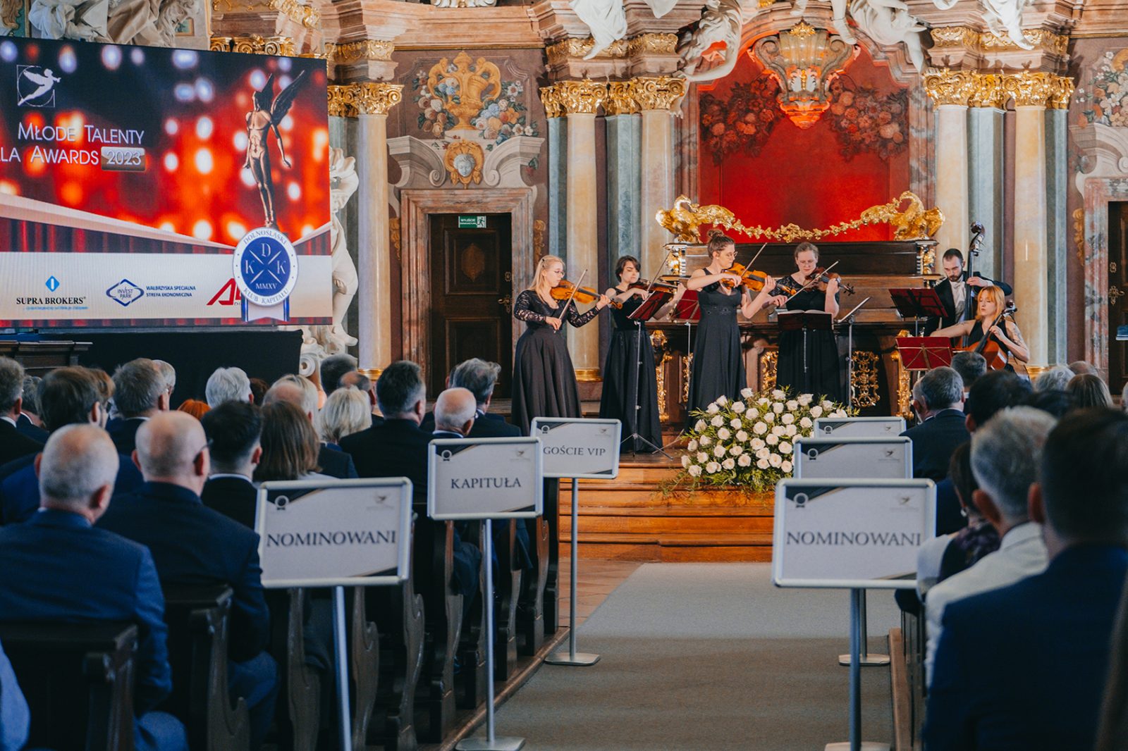 sześcioosobowy zespół muzyczny grający na instrumentach smyczkowych w głebi sali ze zdobieniami na ścianach i suficie