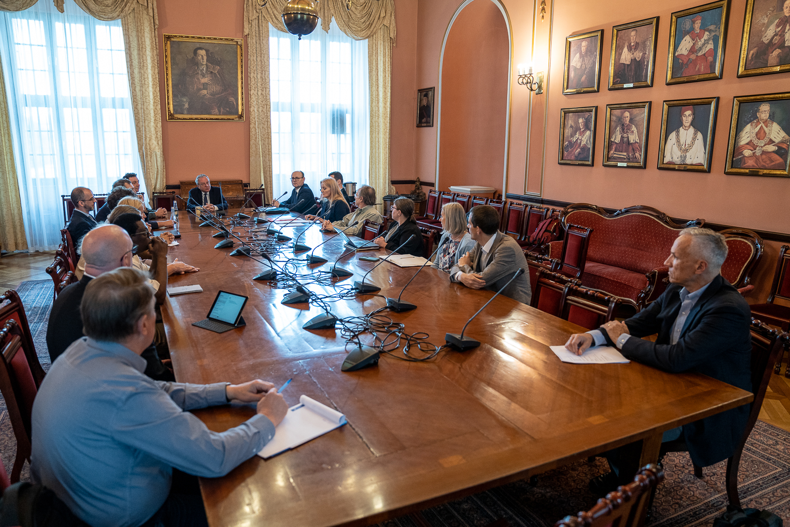 grupa ludzi siedząca przy długim drewnianym zabytkowym stole, na którym stoją mikrofony, siedzą w sali z portretami mężczyzn ubranych w czerwone płaszcze z futrzanymi kołnierzami