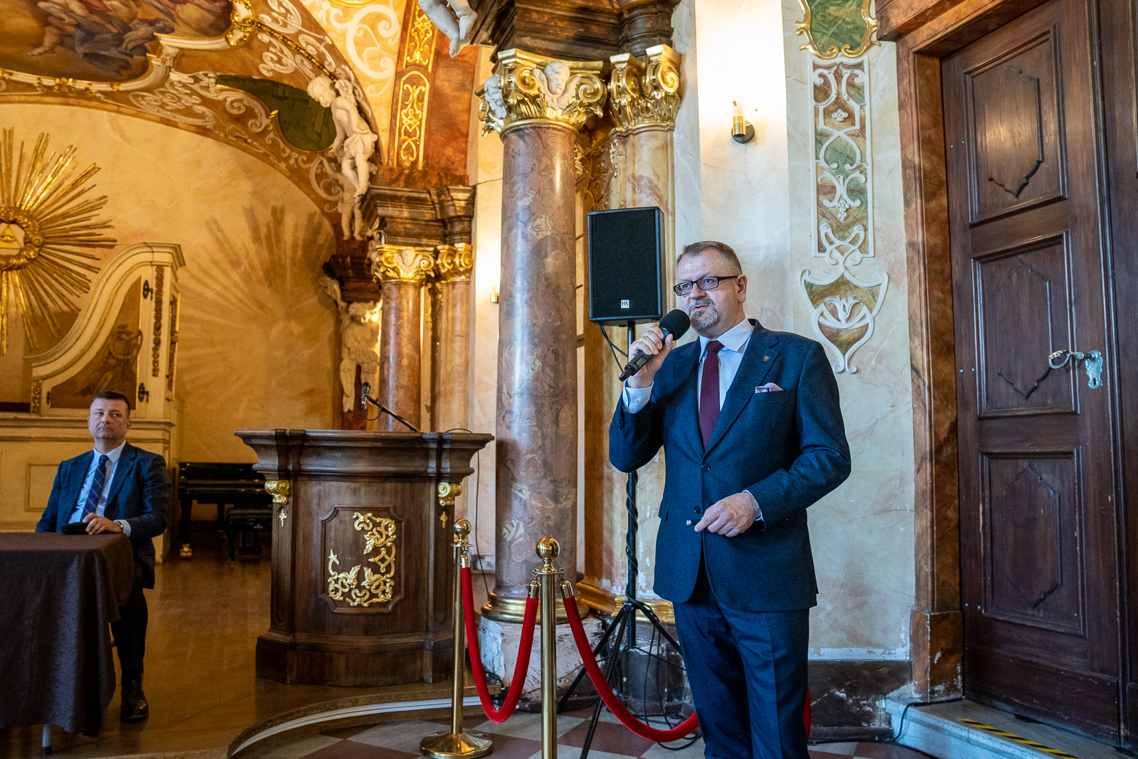 z lewej strony mężczyzna w średnim wieku, siedzący za stołęm w zabytkowej sali, z prawej mężczyzna w granatowym garniturze i fioletowym krawacie trzymający mikrofon