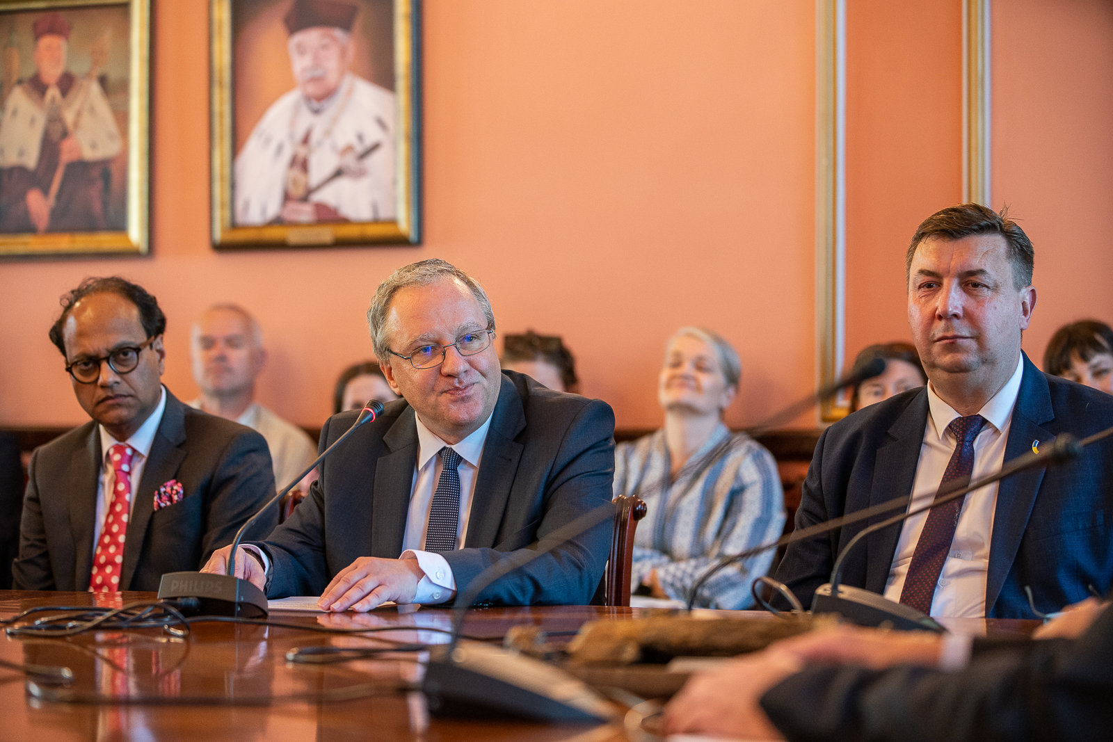 trzech mężczyzn siedzących przy stole na którym stoją mikrofony, mężczyzna z lewej strony ma ciemne włosy, okrągłe okulary, czerwony krawat w białe kropki i ciemną marynarkę, mężczyzna w środku ma siwe włosy okulary, ciemną marynarkę i siwy krawat, mężczyzna z prawej ma ciemne włosy, granatowa marynarkę, białą koszulę i ciemny krawat