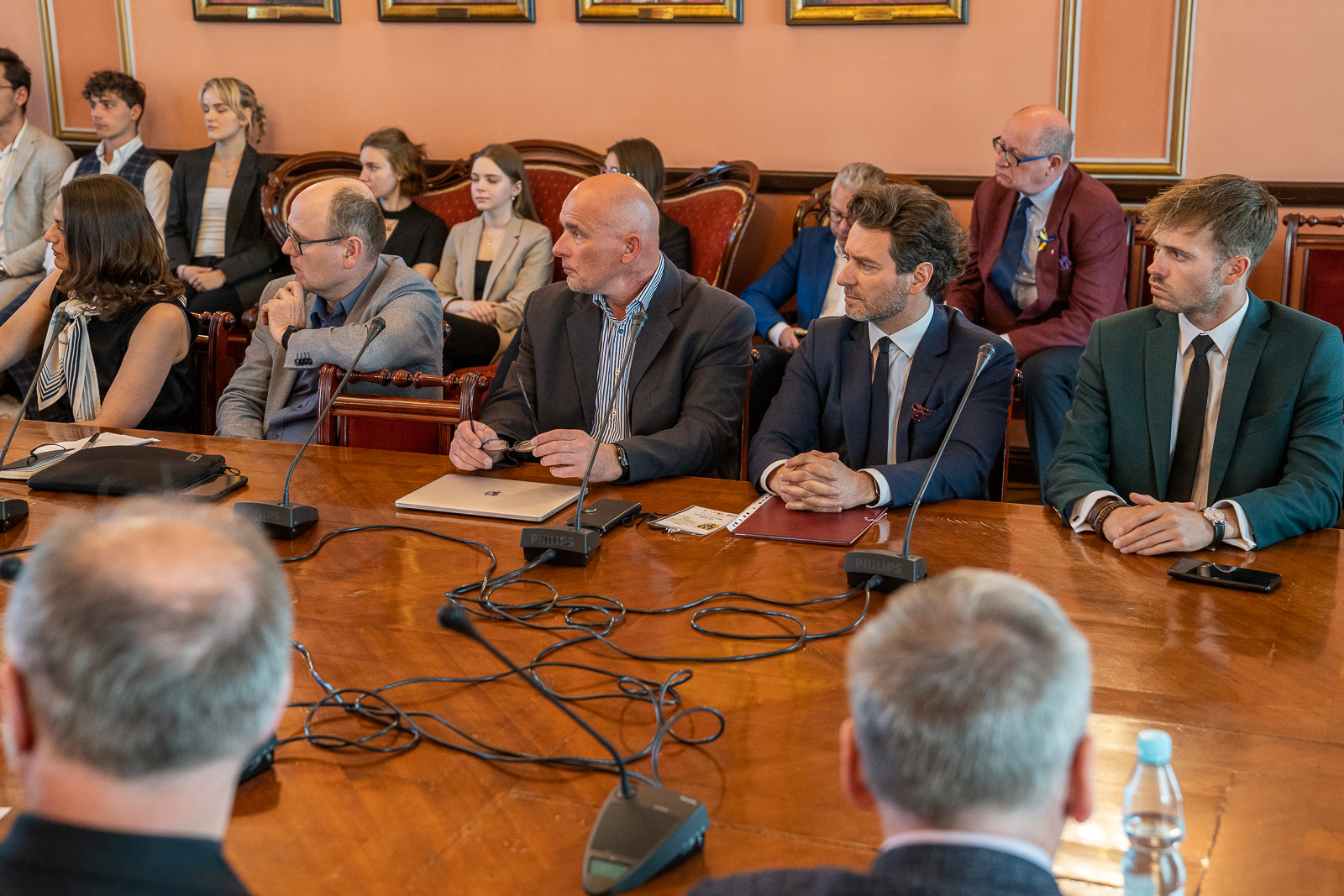 grupa osób siedząca przy dużym drewnianym stole, przed nimi mikrofony, za nimi osoby siedzące na krześle