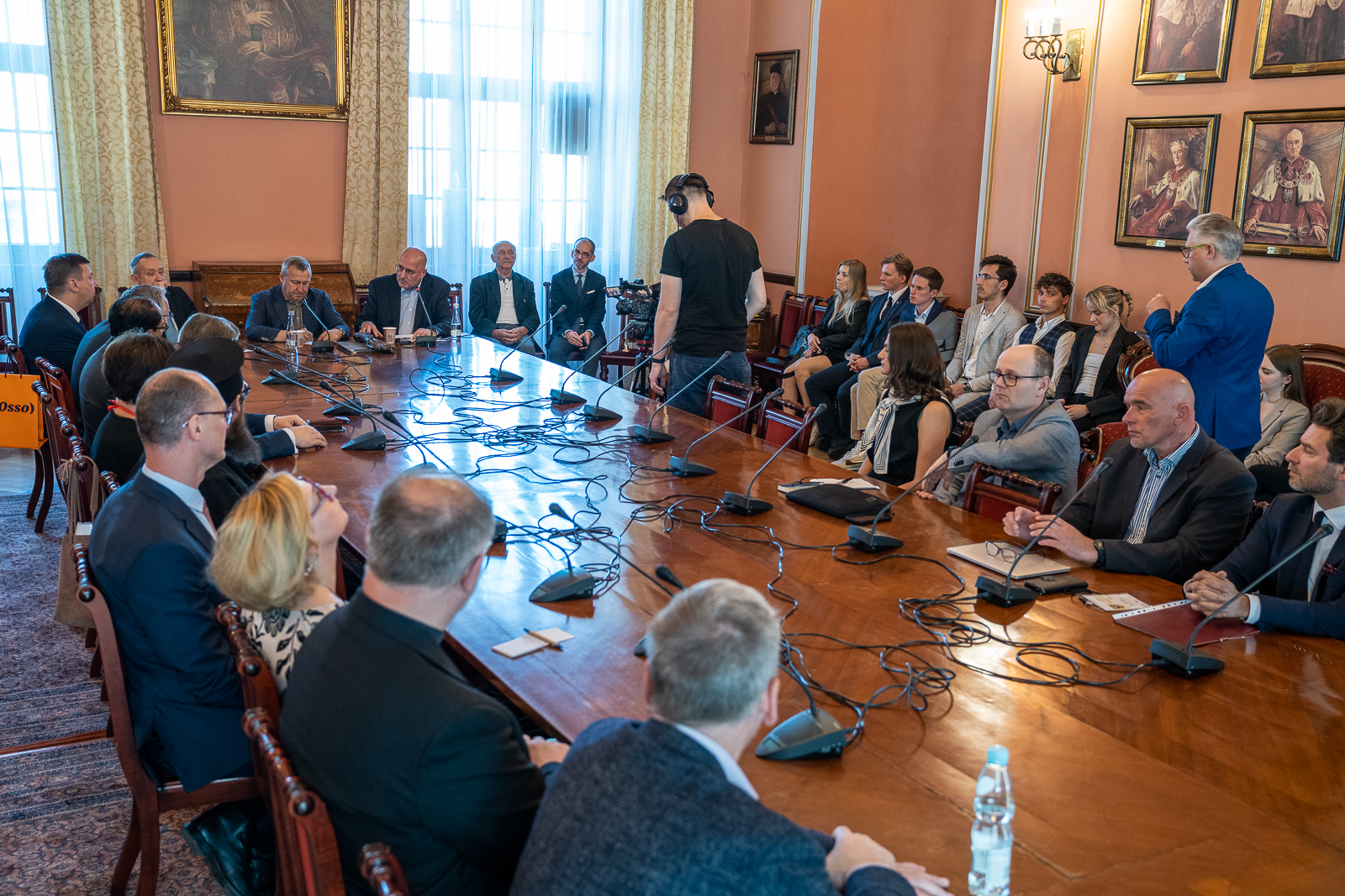 grupa osób siedząca przy dużym brązowym drewnianym stole, na którym stoją mikrofony, w tle ludzie siedzący na krzesłach, na ścianie wiszą obrazy
