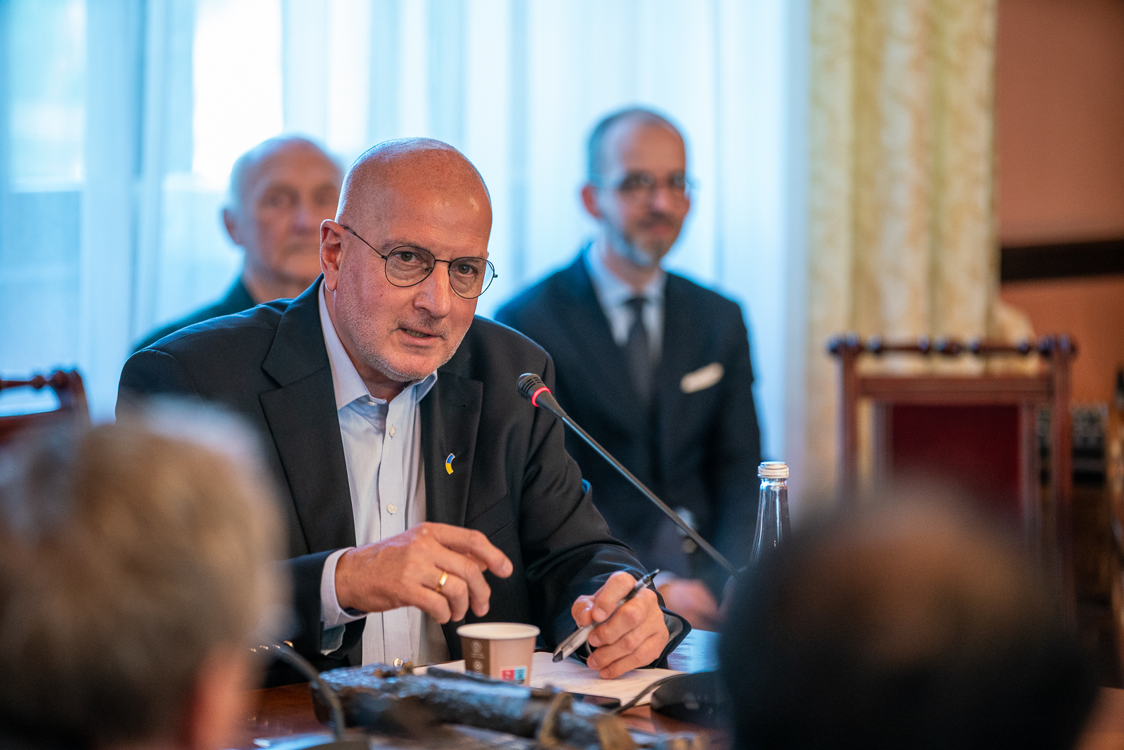 mężczyzna w średnim wieku w okularach z cienkimi oprawkami, jasnoniebieskiej koszuli i ciemnej marynarce siedzący przy stole przed nim mikrofon, zanim dwóch mężczyzn