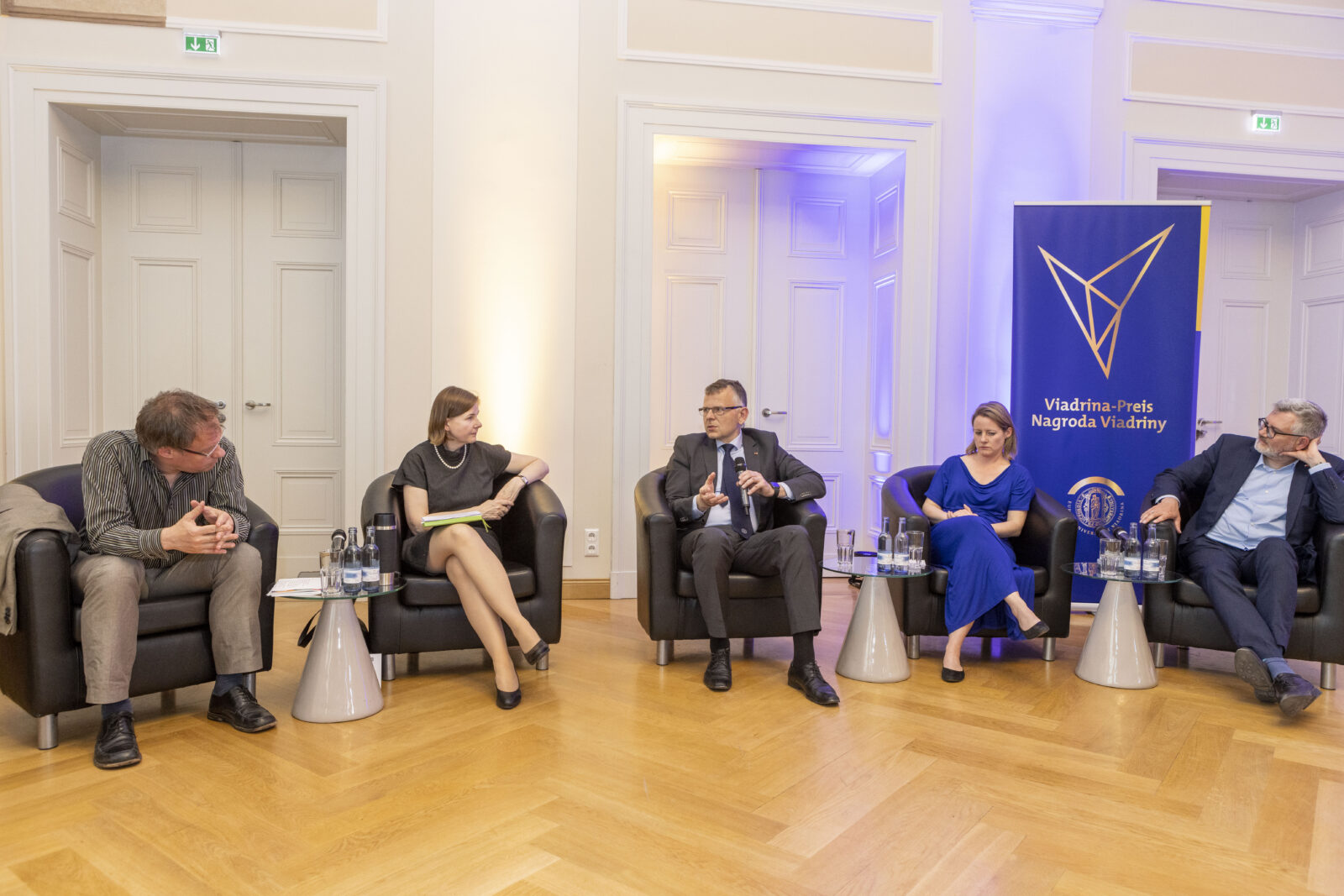 pięć osób w średnim wieku siedzi w fotelach w sali z parkietem pod białą ścianą z trojgiem drzwi