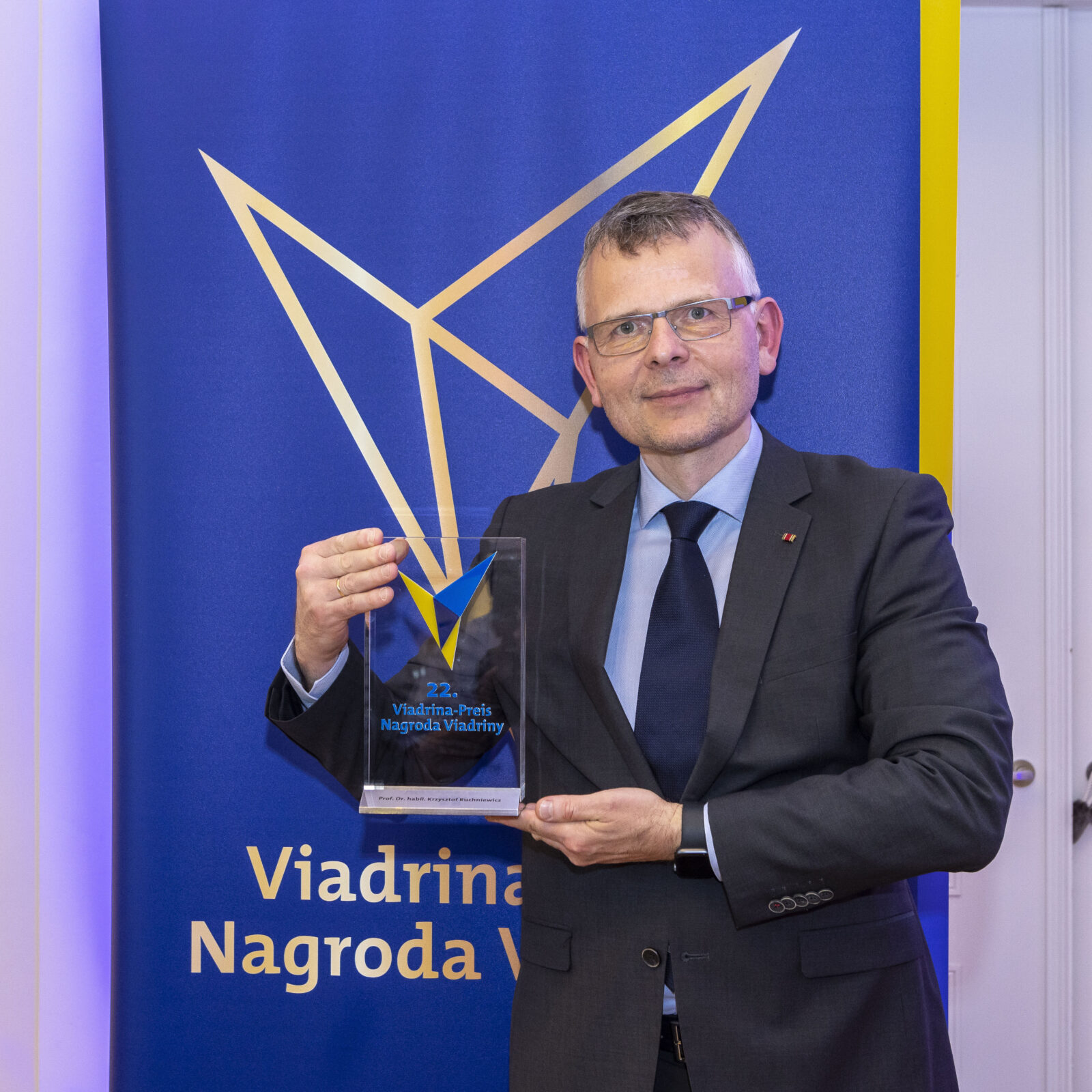 Mężczyzna w średnim wieku w okularach, garniturze, niebieskiej koszuli z krawatem trzyma w rękach prostokątna, przezroczystą statuetkę