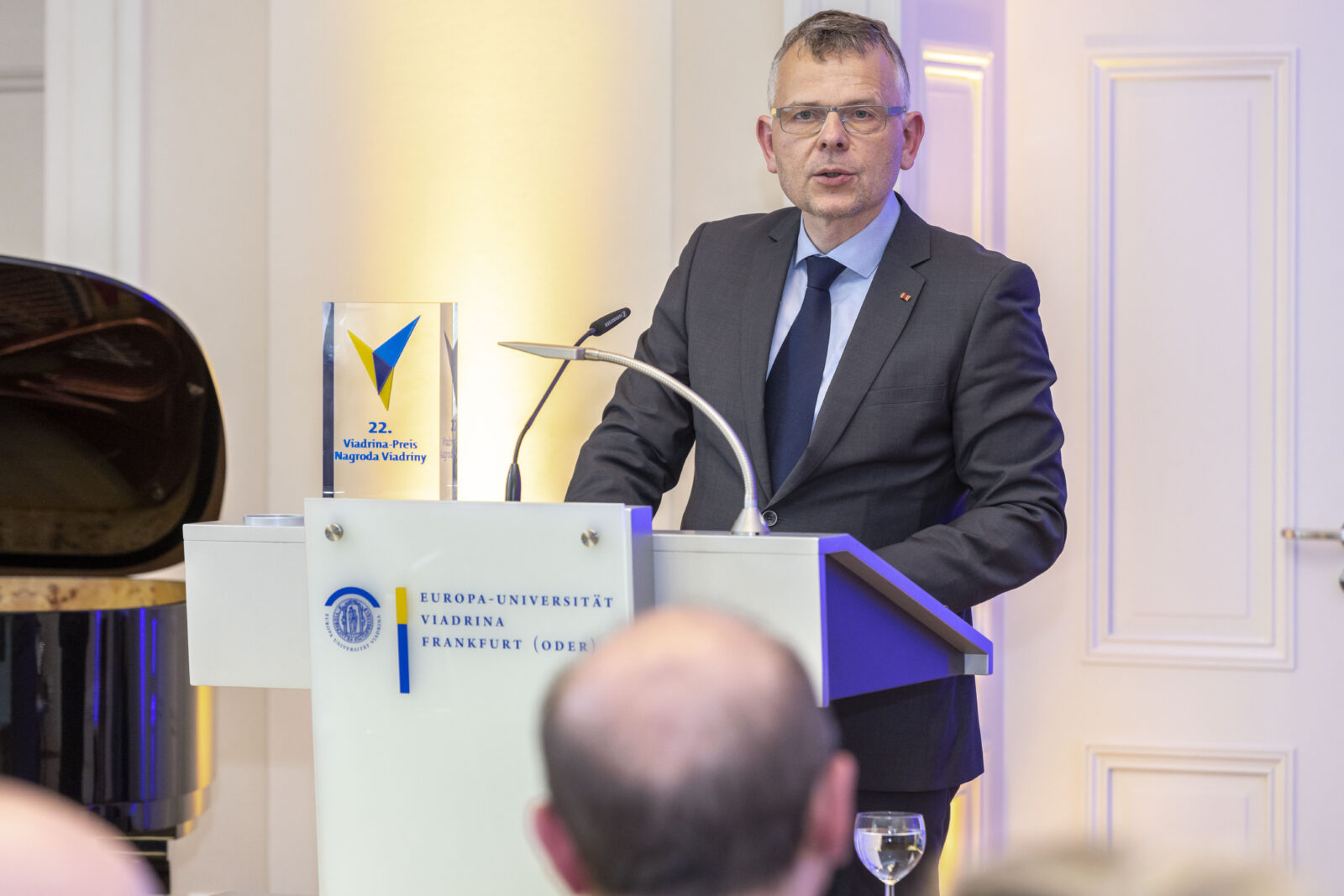 Mężczyzna w średnim wieku w okularach, garniturze, niebieskiej koszuli z krawatem stoi przy białym pulpicie z mikrofonem