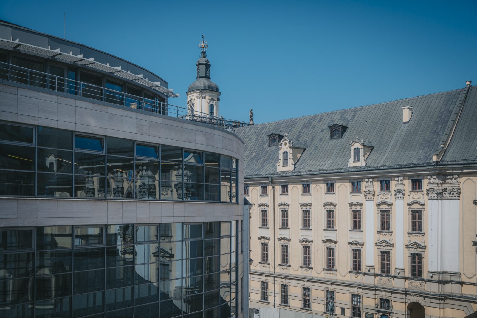dwa duże budynki, po lewej stronie nowoczesny, z przeszklonymi ścianami, po prawej stronie, w głębi, budynek znacznie starszy, ze spadzistym szarym dachem, wieżą i wieloma ozdobnymi oknami