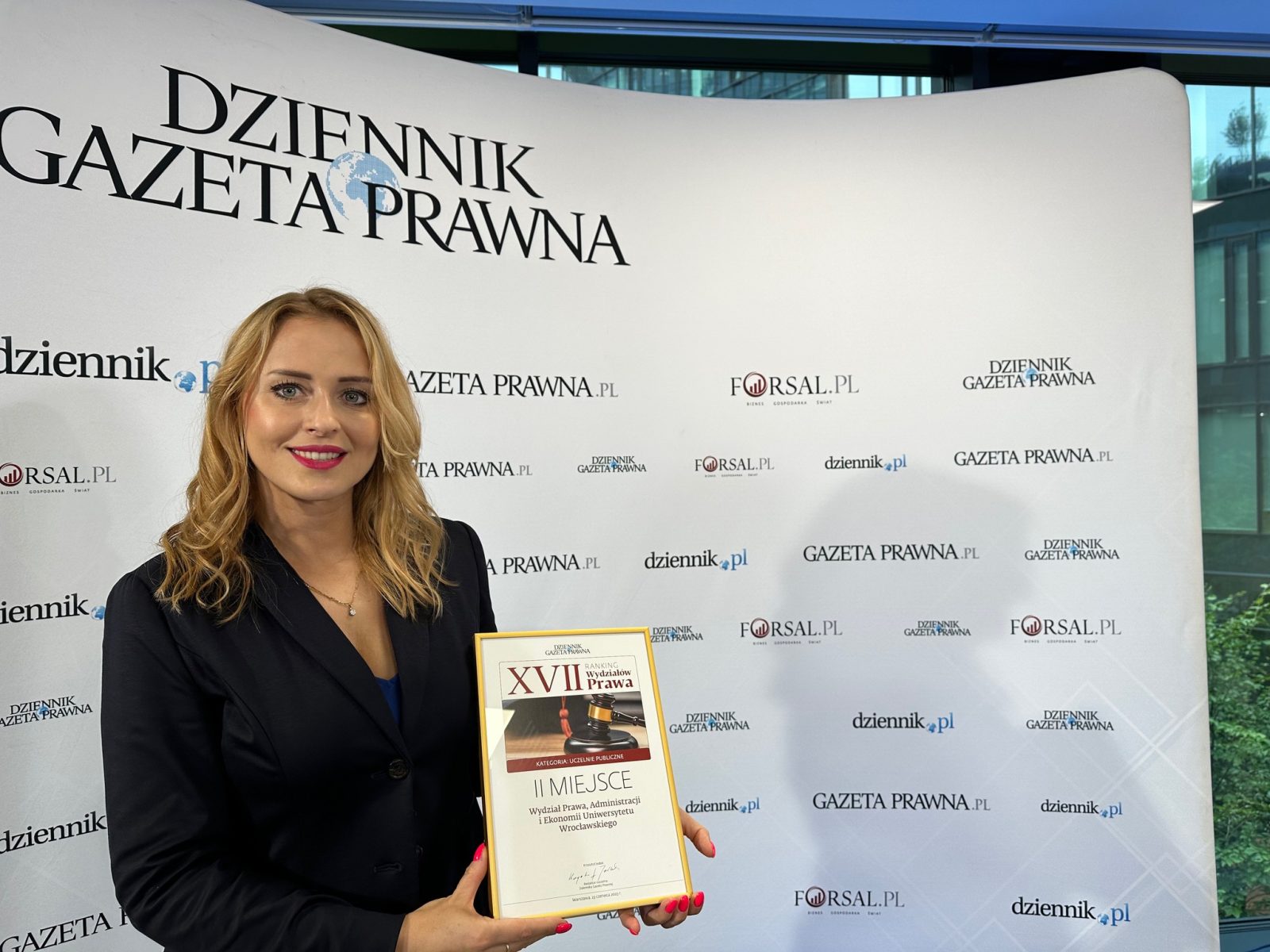 młoda kobieta o długich jasnych włosach, w czarnej marynarce, trzymająca w rękach dyplom, stoi na tle ściany z napisami dziennik.pl, gazeta.pl, foksal.pl