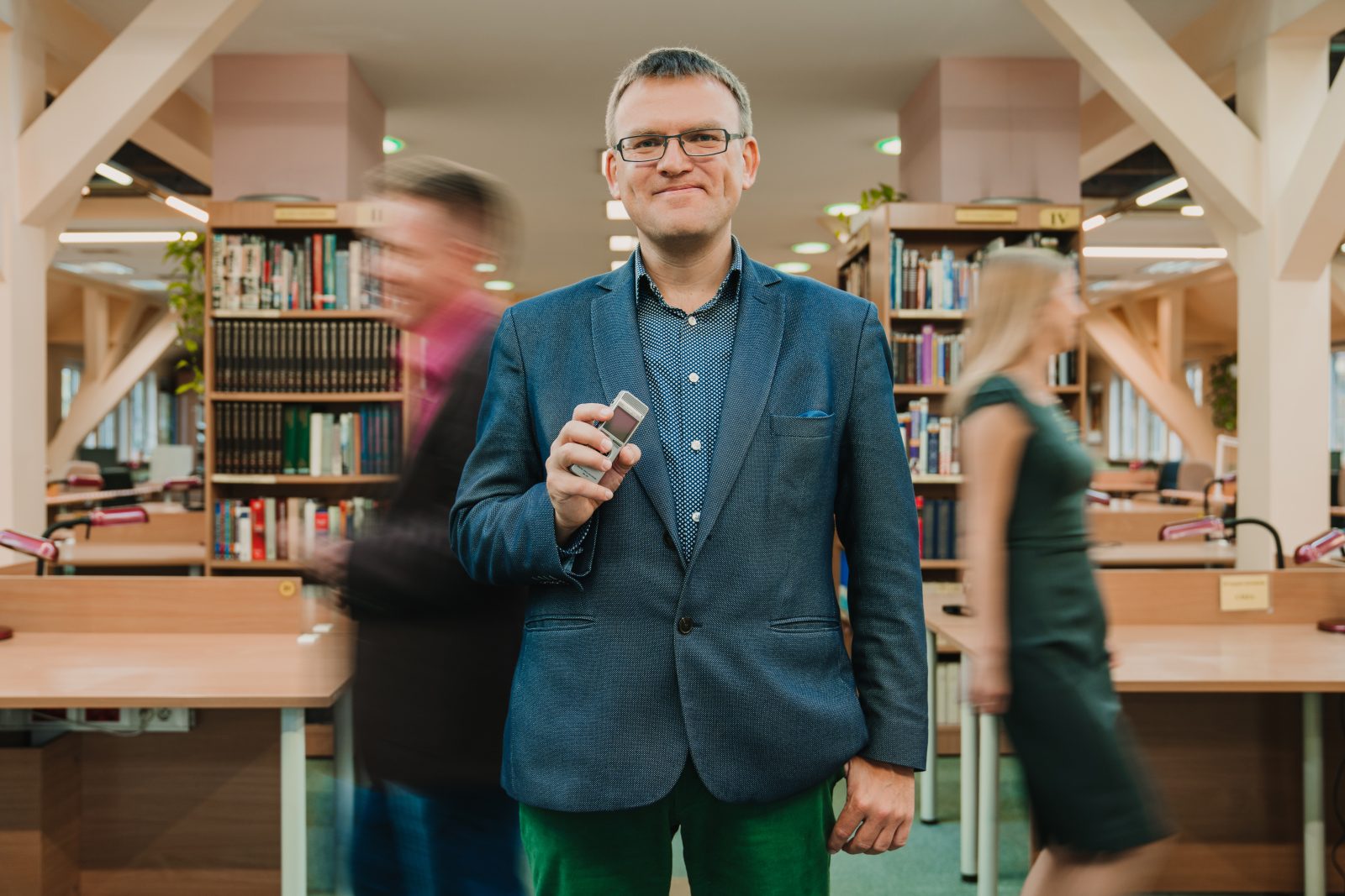 Mężczyzna w średnim wieku w garniturze trzyma w dłoni mały dyktafon, w tle dwoje ludzi w ruchu i regały z książkami