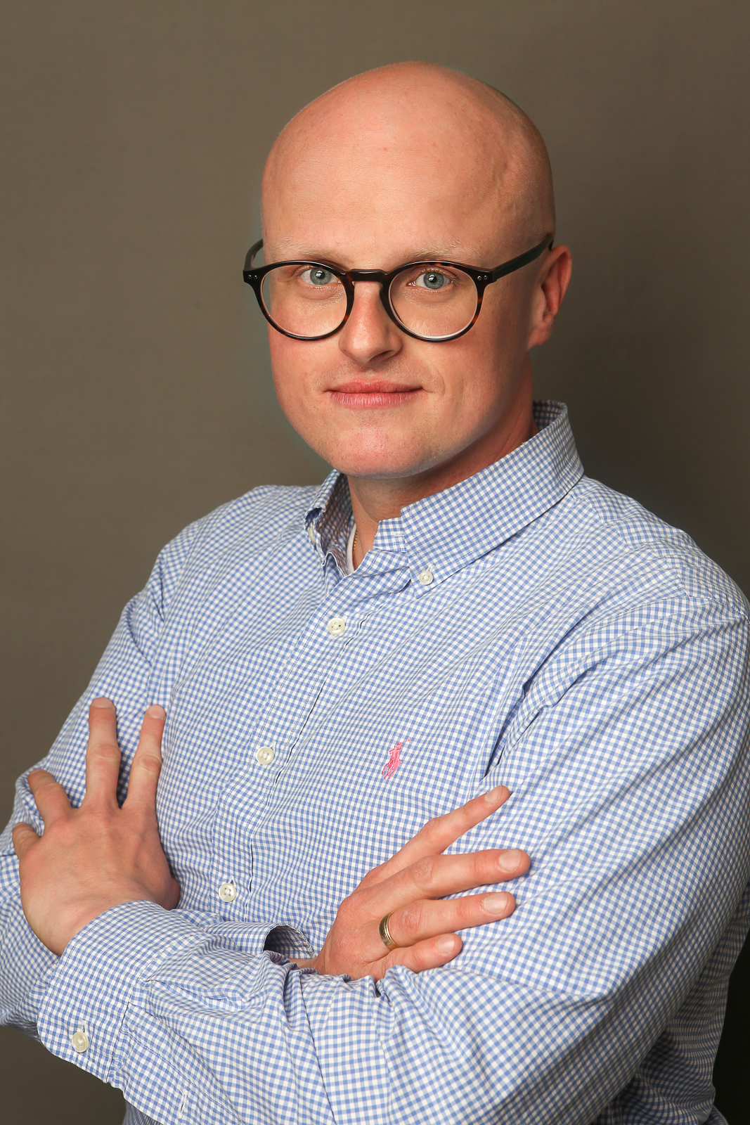 młody mężczyzna w okularach w koszuli w drobną niebieską kratkę, stoi z założonymi rękoma