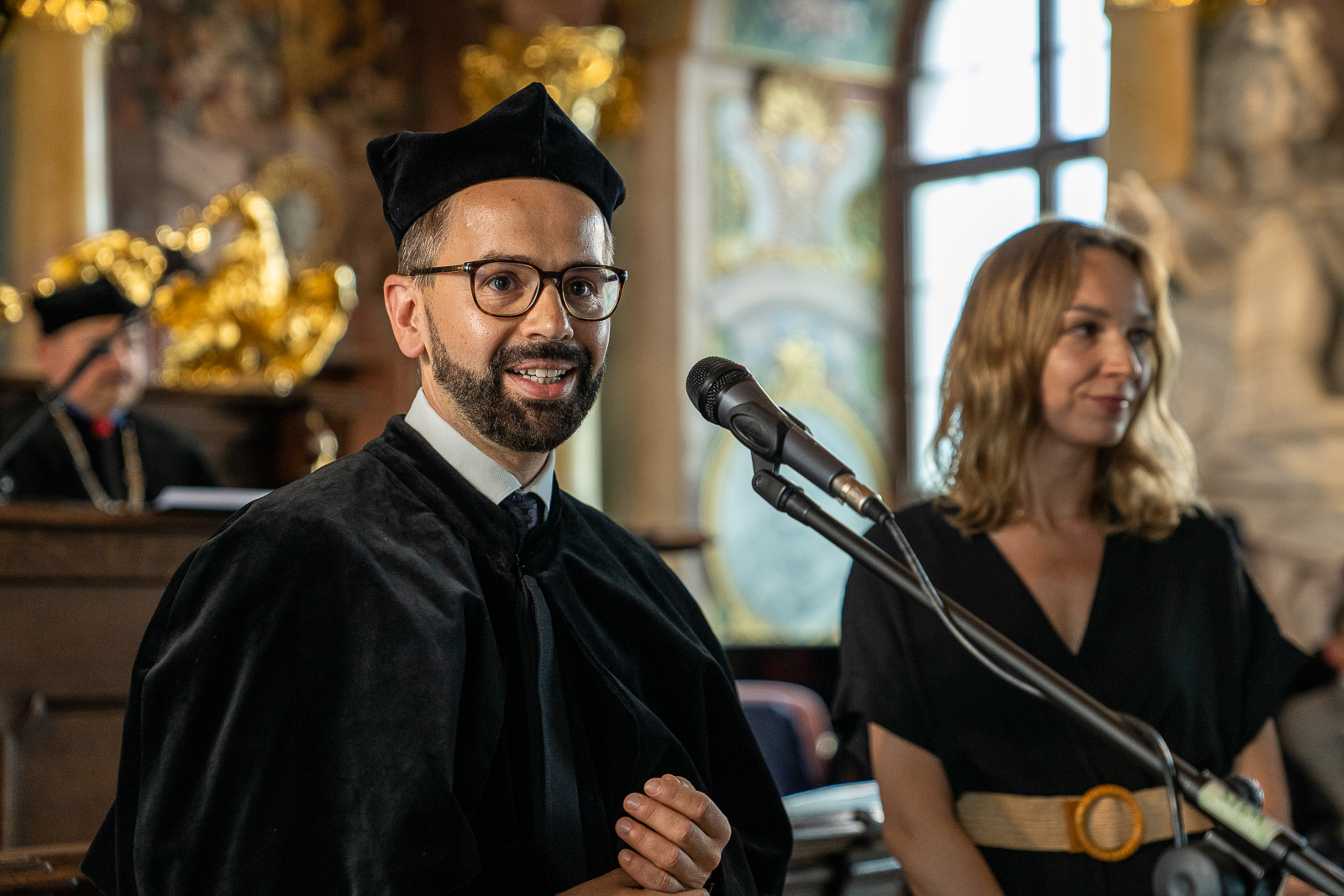 mężczyzna w czarnym płaszczu i czapce i okularach, z brodą, stojący przed mikrofonem, obok kobieta w czarnej sukience, stoją w zabytkowej sali o ścianach pokrytych malowidłami i rzeźbami