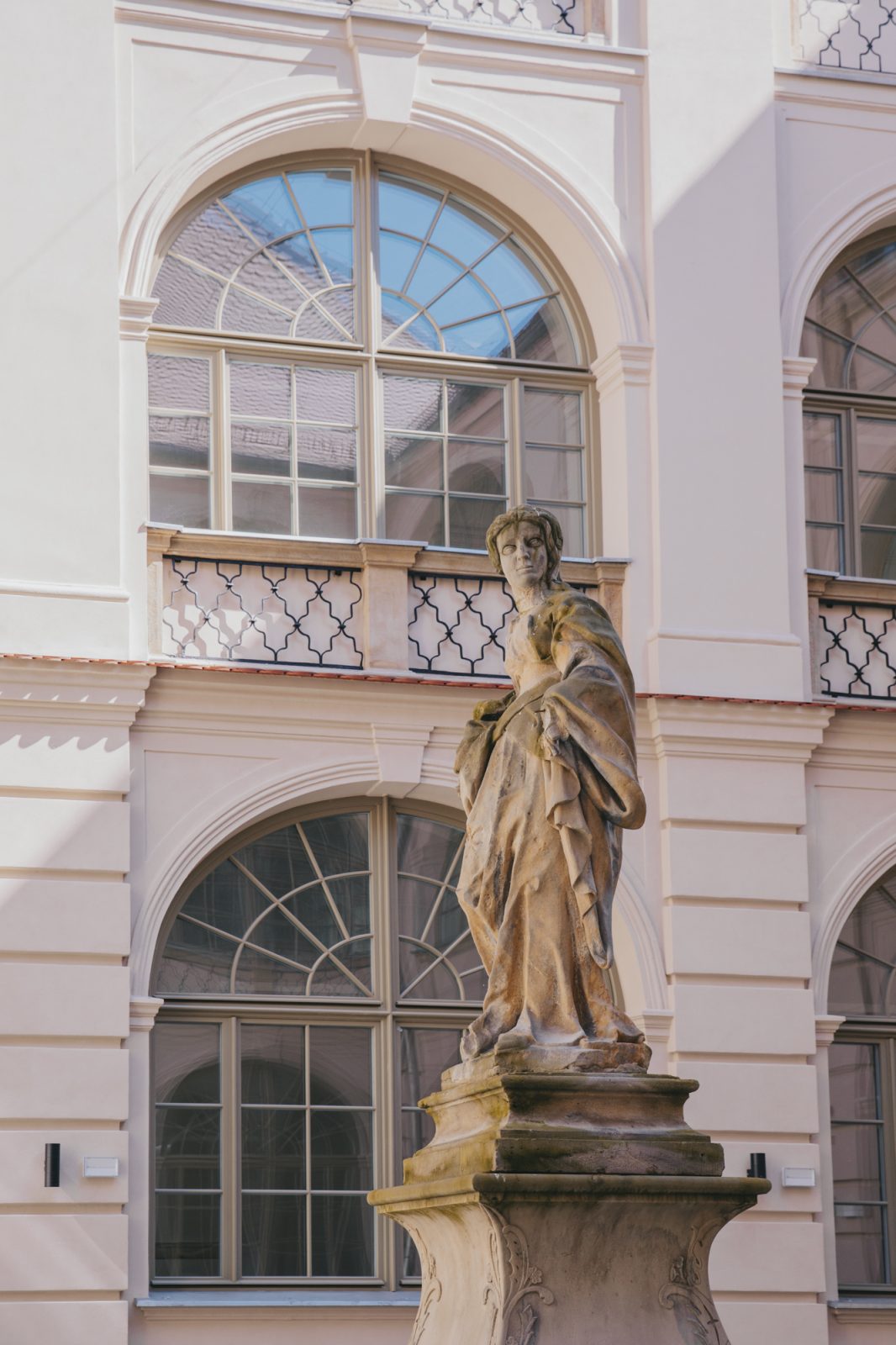 zabytkowa figura, w tle zabytkowy budynek z półokrągłymi oknami