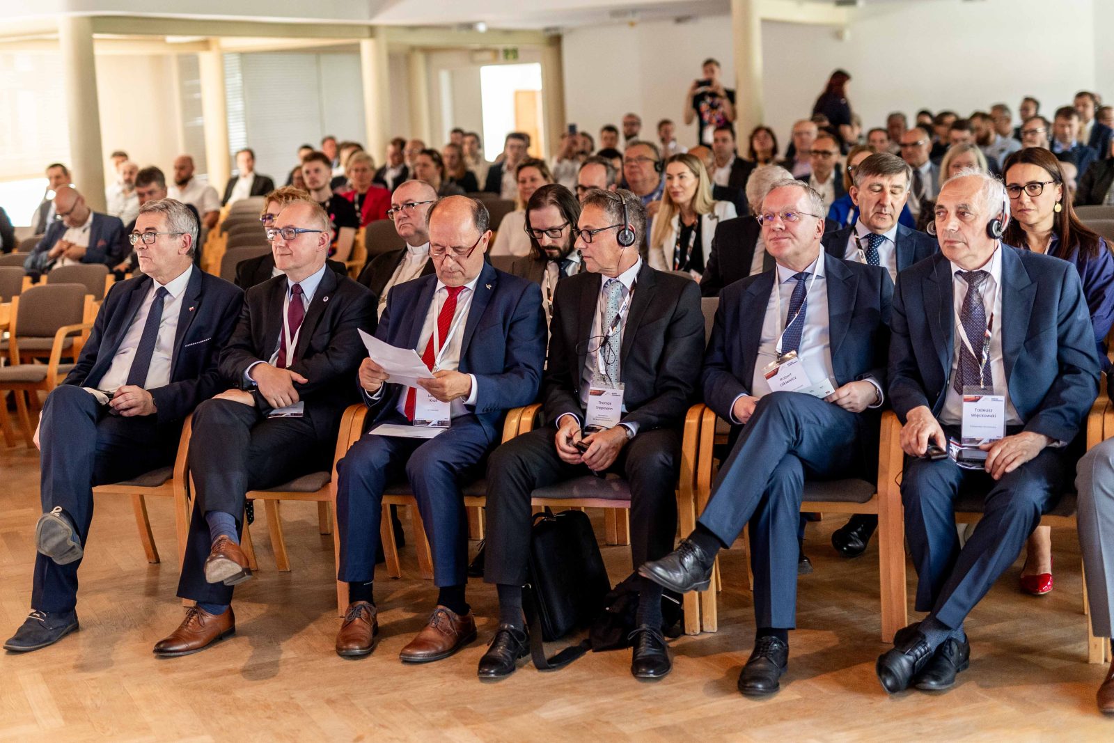grupa osób siedząca na drewnianych krzesłach, na pierwszym planie mężczyźni w średnim wieku w ciemnych garniturach, białych koszulach i krawatach, dwóch z nich ze słuchawkami na uszach