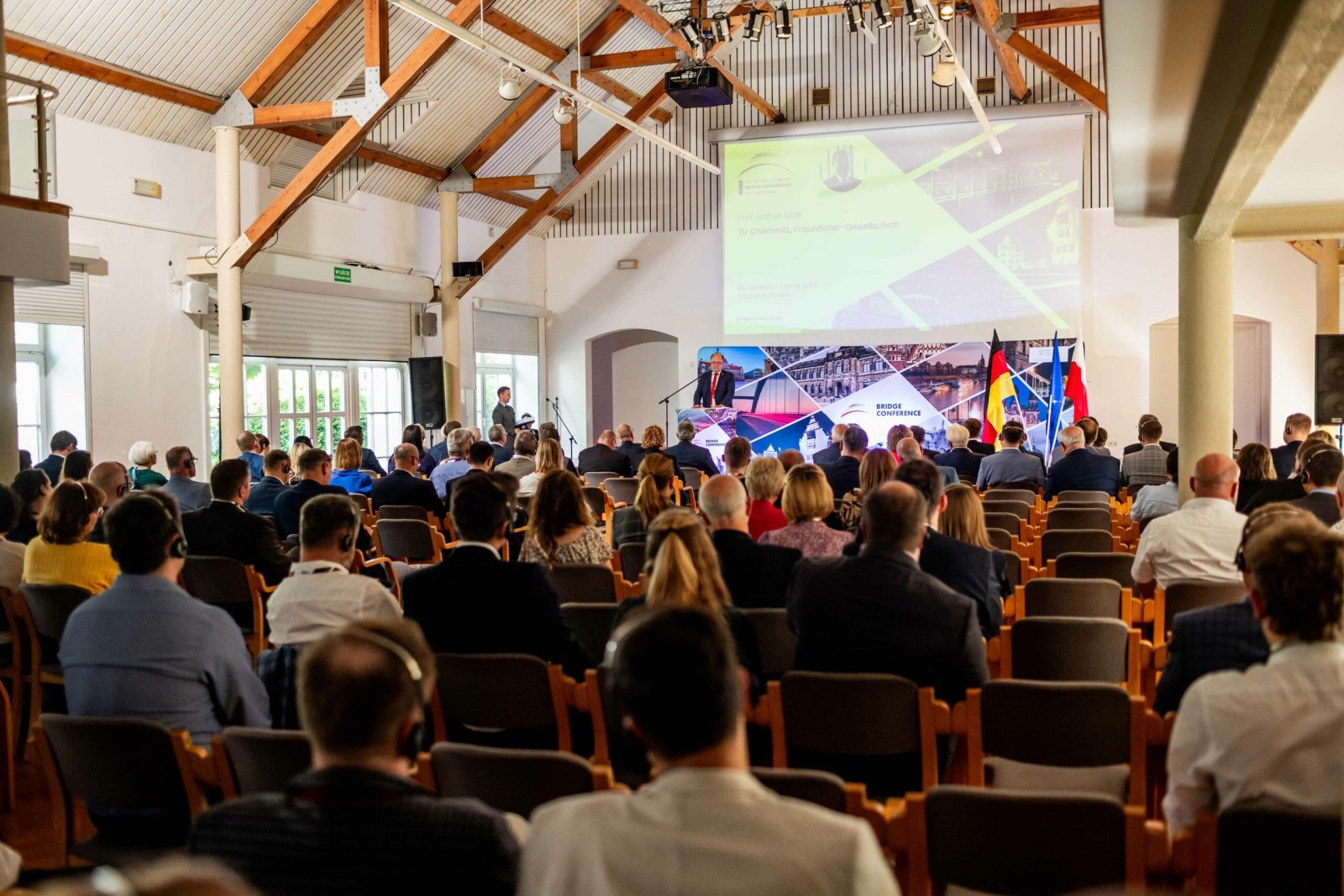 grupa osób siedząca w sali na drewnianych krzesłach, widoczna od tyłu, z przodu ekran z kolorowymi zdjęciami, na podeście mężczyzna w garniturze