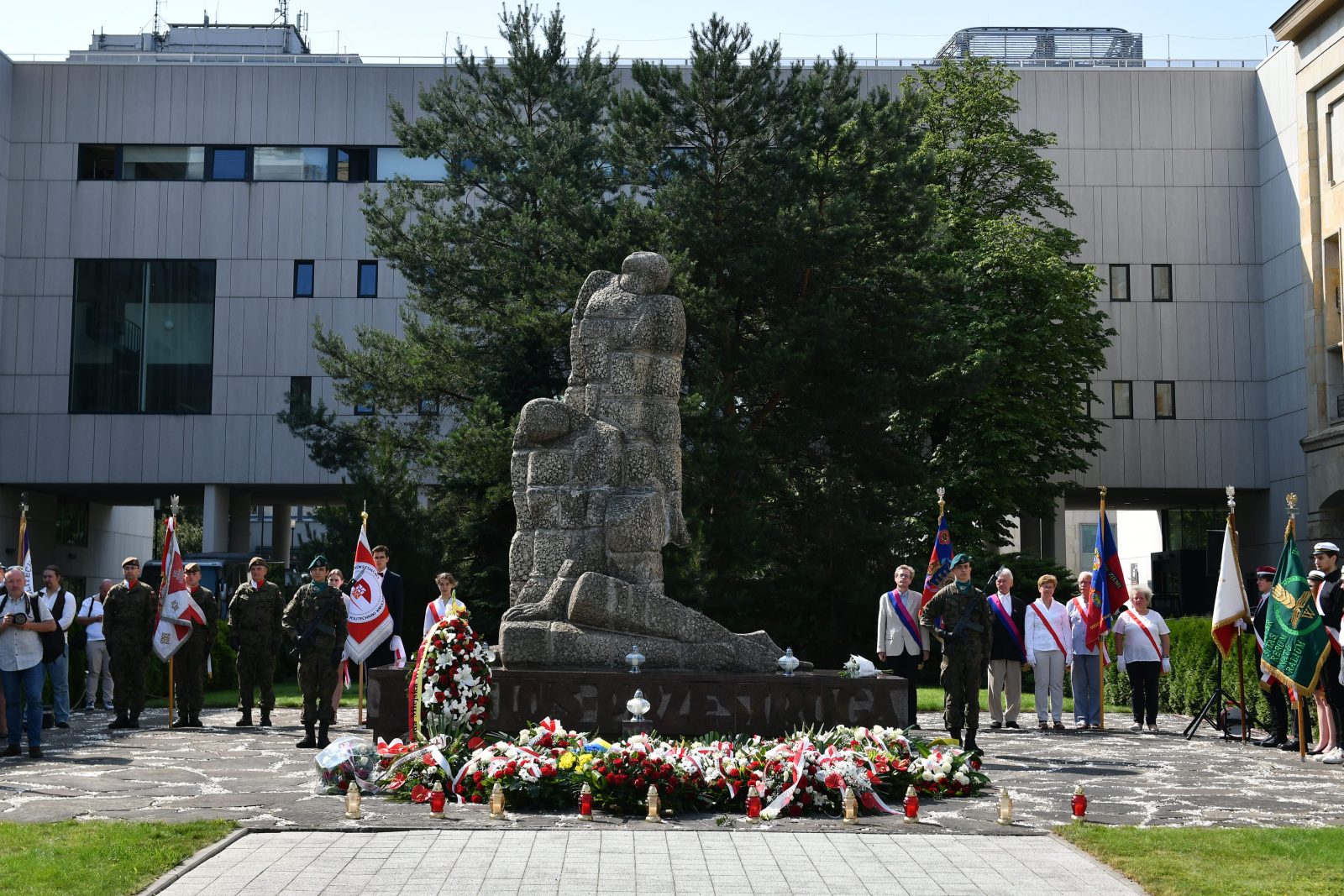 duży kamienny pomnik przedstawiający rzeźbę dwóch siwych postaci, jedna w pozycji leżącej a druga stojącej. Po obu stronach pomnika stoją osoby z dużymi chorągwiami, przed pomnikiem wieńce z kwiatów, za pomnikiem stoi duży siwy budynek