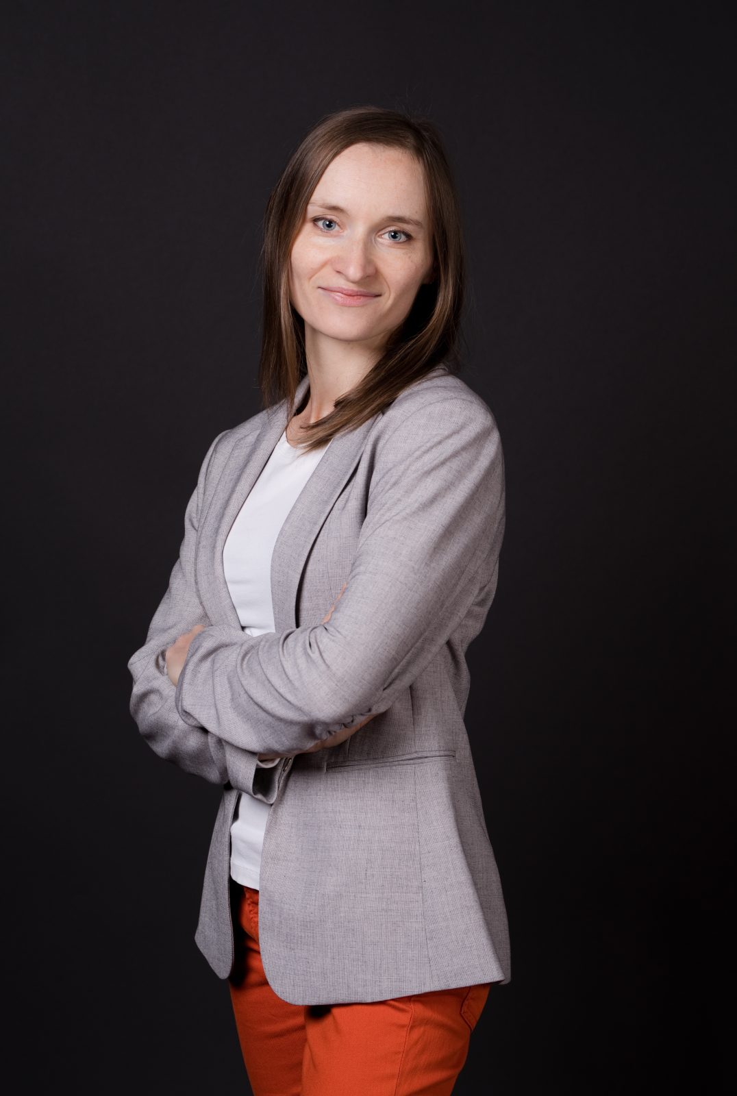 Portret kobiety ukazanej do połowy uda. Szczupła kobieta z brązowymi włosami do ramion. Ubrana jest w białą koszulkę, szarą marynarkę i czerwone spodnie.