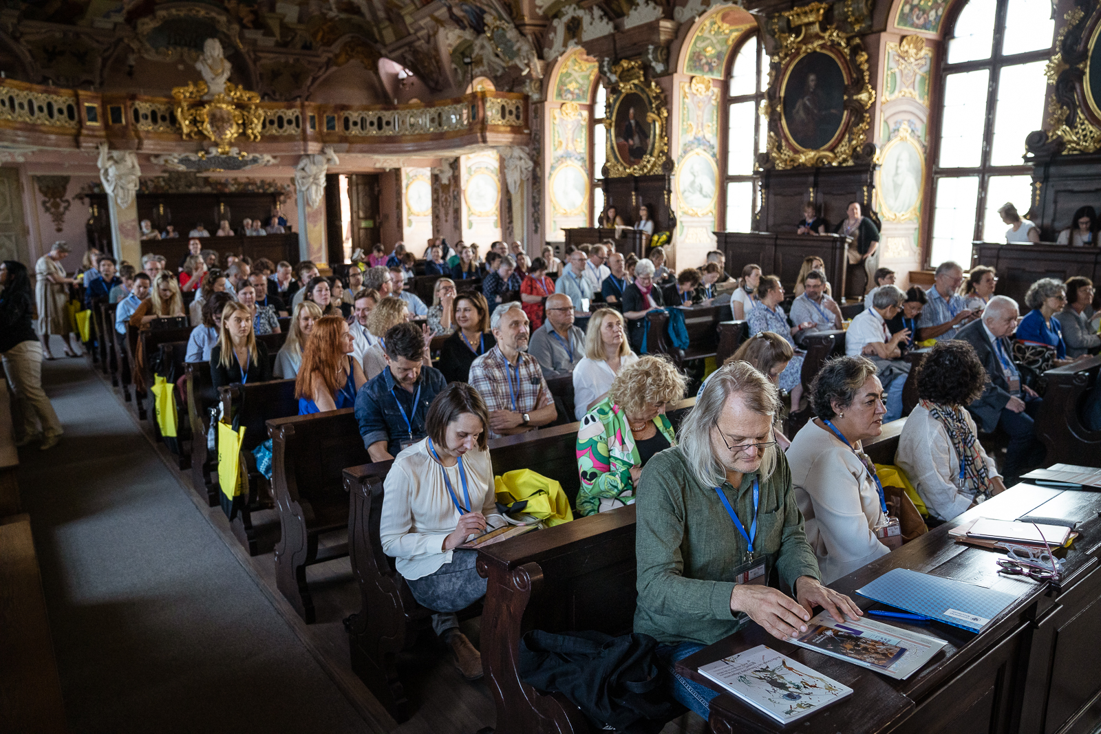 Duża sala ze zdobieniami na ścianach, portretami w złotych ramach, dużymi oknami, wypełniona siedzącymi w ławkach ludźmi