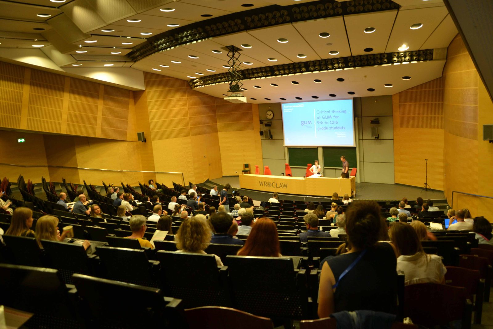 duża sala konferencyjna widziana z górnych rzędów krzeseł. Na widowni, na krzesłach siedzi wiele osób, a u dołu, na scenie, za pulpitem z napisem Wrocław stoją dwie osoby. Nad nimi wisi rozwinięty ekran rozświetlony niebieskim obrazem z rzutnika.