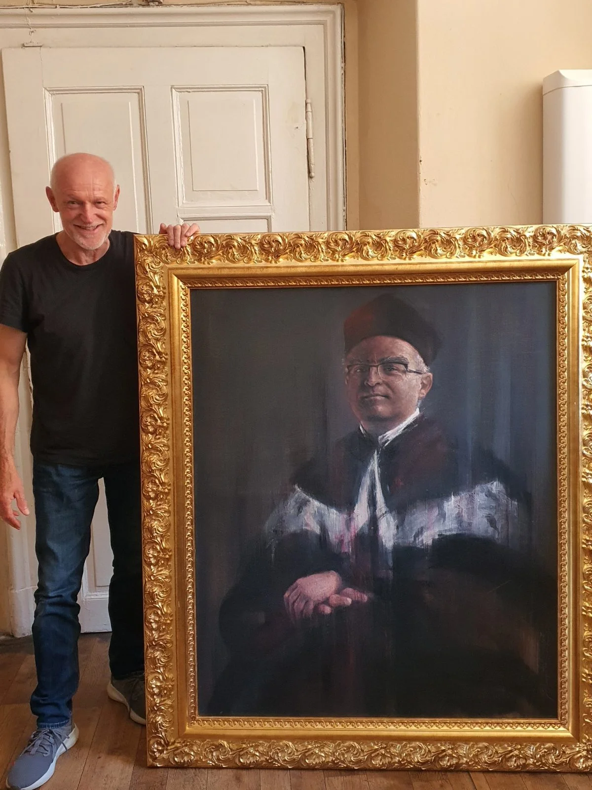 Mężczyzna w średnim wieku ubrany na czarno trzyma portret rektorski oprawiony w złotą ramę.
