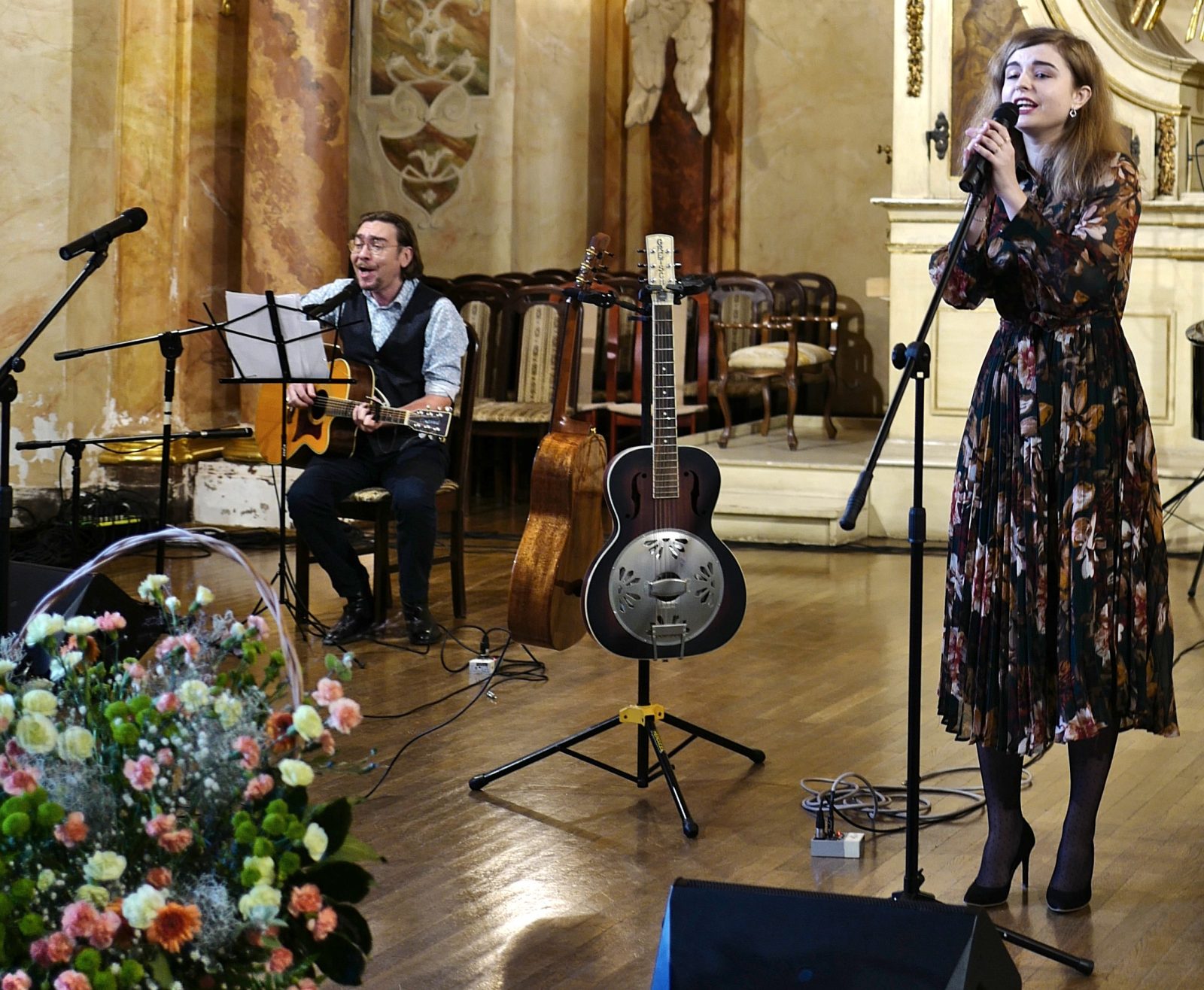 Po lewej stronie mężczyzna w średnim wieku siedzi na krześle z oparta na kolanach gitarą, po prawej stronie kobieta w średnim wieku stoi przy mikrofonie na statywie. Pbok nich ustawiony jest duży bukiet kwiatów oraz dwie gitary na podstawkach.