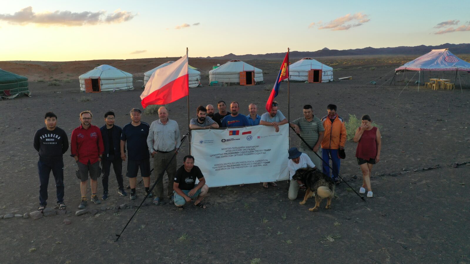 Kilkanaście osób stoi lub kuca za i po bokach białego baneru ze słabo widocznym napisem. Bo bokach baneru są zatknięte flagi Polski i Mongolii. W tle stoi kilka namiotów-jurt, na horyzoncie widoczne zarysy gór.
