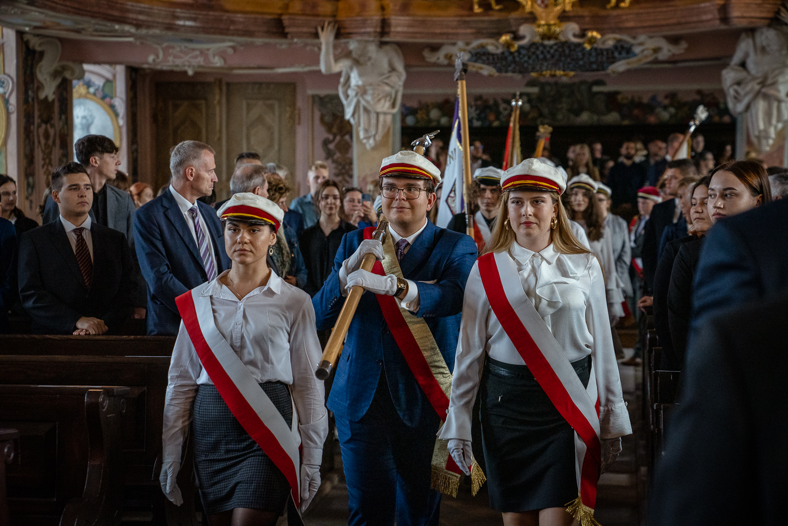 Młodzi ludzie przepasani biało-czerwonymi szarfami niosą sztandary w sali wypełnionej ludźmi