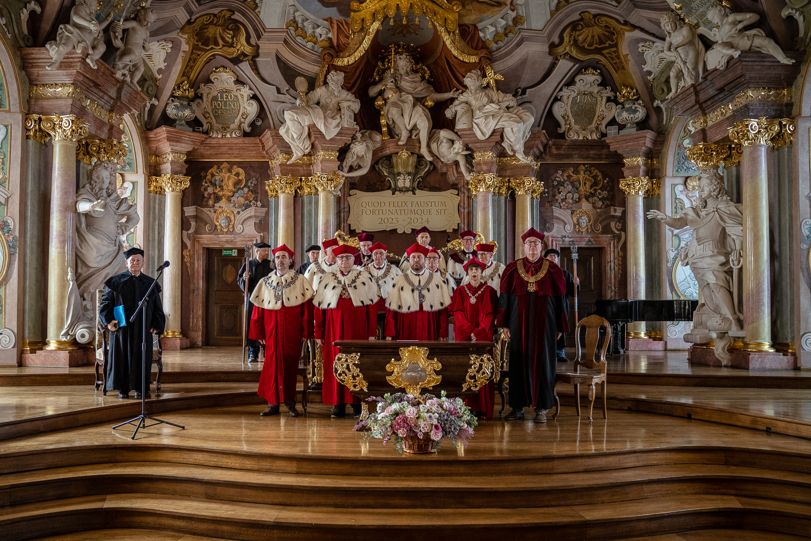Grupa kilkunastu osób w strojach rektorskich stoi na podium pod ścianą z ozdobnymi kolumnami i wieloma białymi rzeźbami