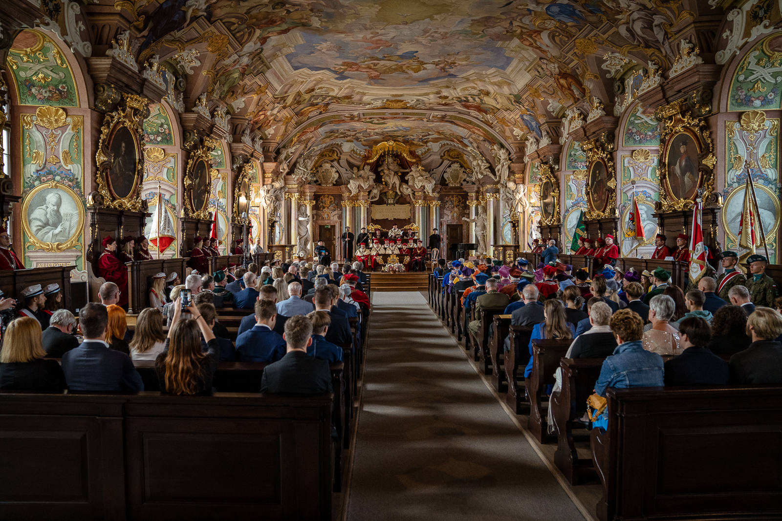 Duża sala ze ścianami i sufitem ozdobionymi obrazami, płaskorzeźbami i freskami. W dwóch kolumnach ławek siedzą ludzie widziani od tyłu.