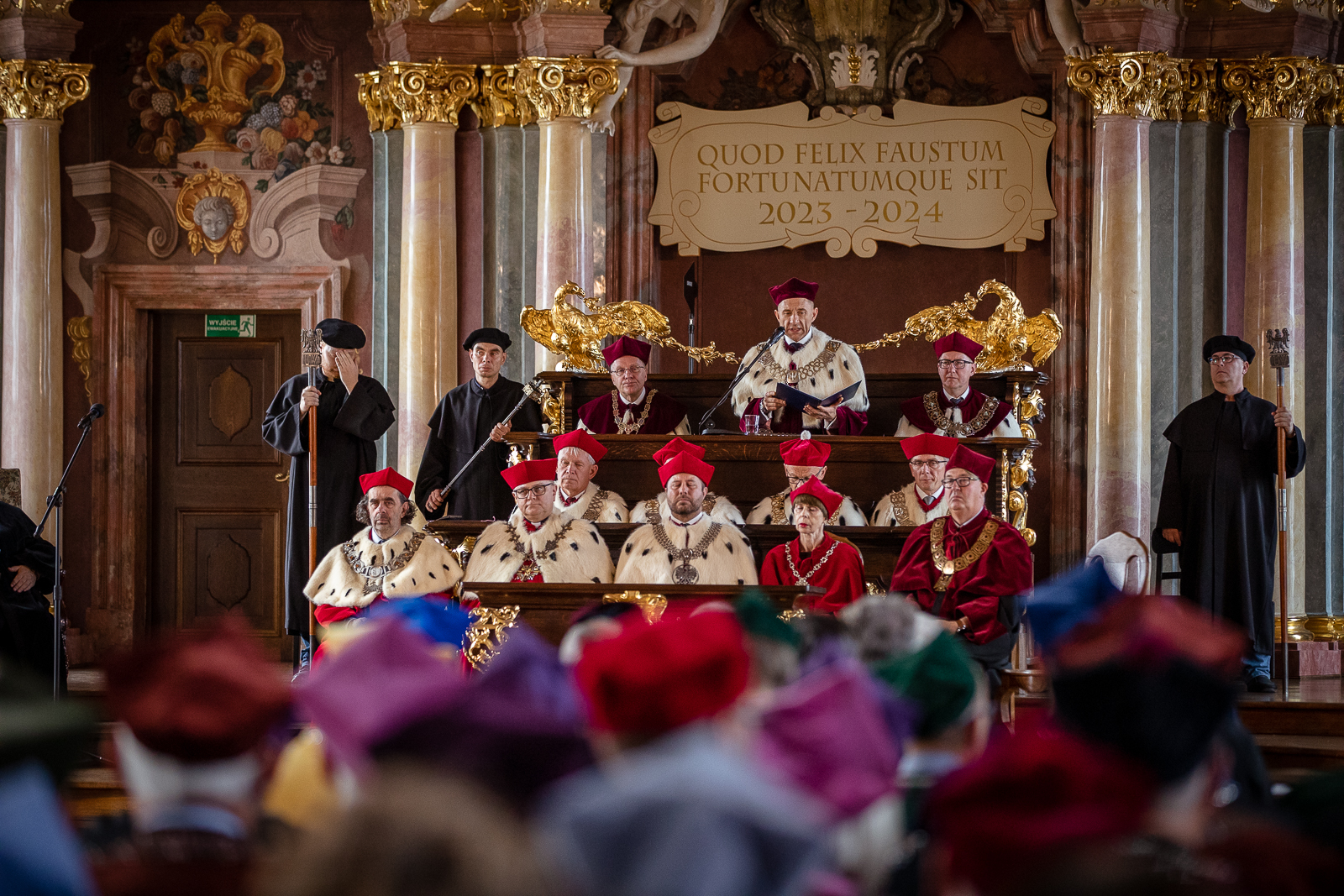 Grupa kilkunastu osób w togach i biretach siedzących w trzech rzędach ławek ze złotymi zdobieniami pod ścianą z kolumnami