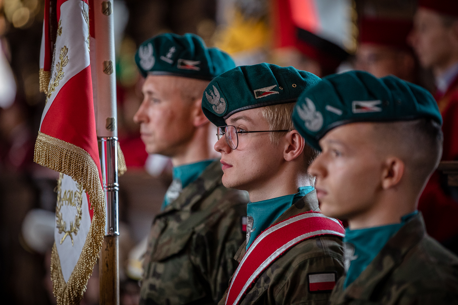 Trzej młodzi mężczyźni widoczni z profilu, na głowach maja zielone berety z orłem. Przed nimi widoczny częściowo sztandar ze złotymi frędzlami.