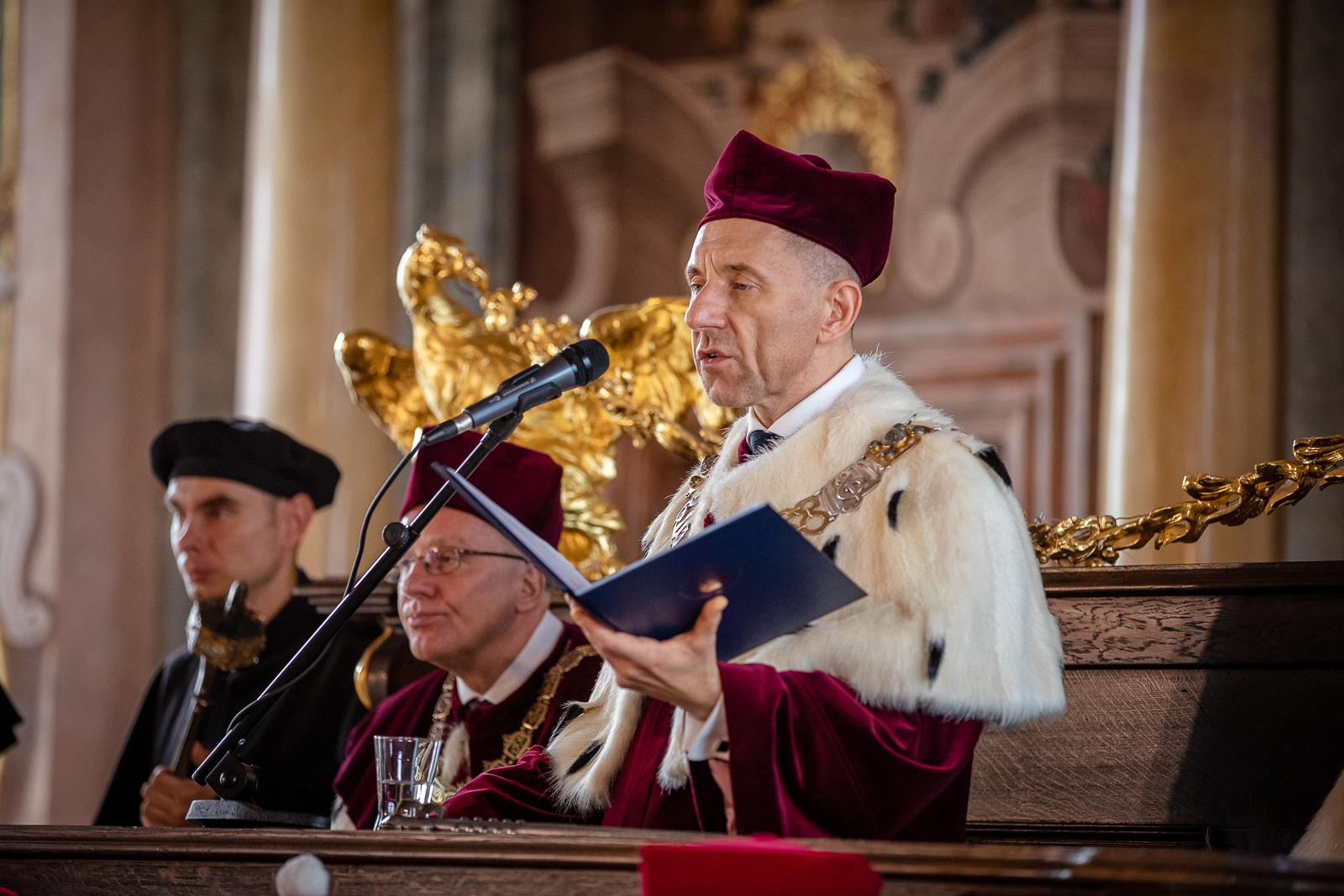 Widoczny do pasa mężczyzna w purpurowym birecie i todze rektorskiej zwrócony twarzą do mikrofonu na statywie. W dłoni trzyma otwartą okładkę na dokumenty. Za nim widać innego mężczyznę w podobnym stroju oraz drugiego w czarnym stroju.