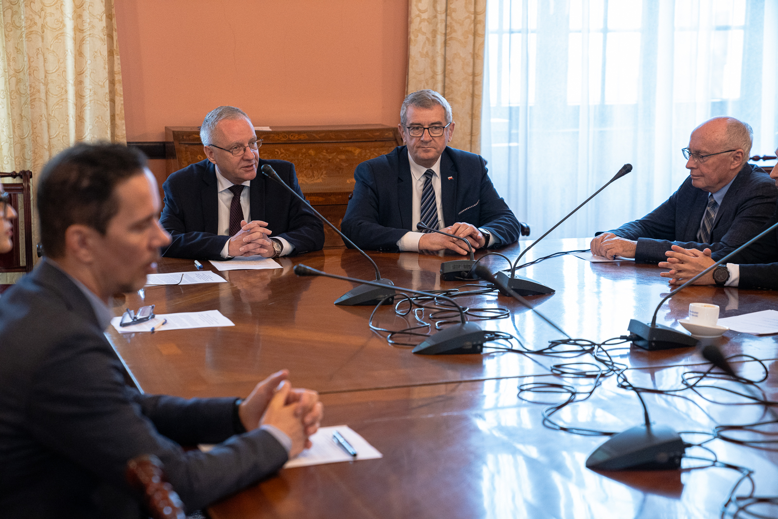 grupa czterech mężczyzn w garniturach i krawatach siedząca przy dużym stole , na którym stoją mikrofony