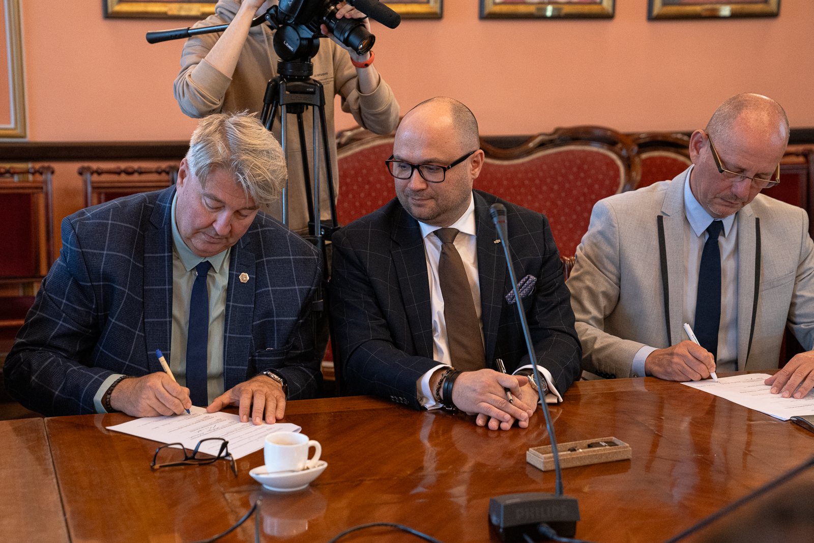 trzech mężczyzn w średnim wieku siedzący przy dużym stole , na którym stoi mikrofon, podpisują dokumenty