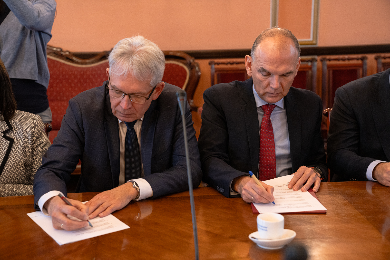 dwóch starszych mężczyzn siedzący przy dużym stole , na którym stoi mikrofon, podpisują dokumenty