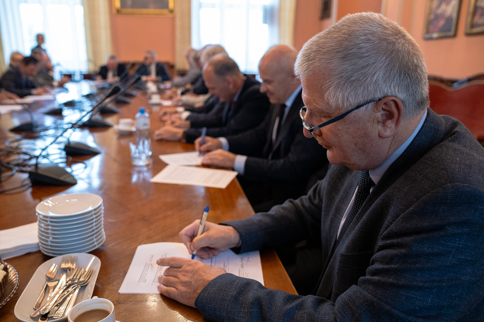 grupa mężczyzn w starszym wieku siedząca przy dużym stole , na którym stoją mikrofony, dwaj mężczyźni z przodu podpisują dokumenty