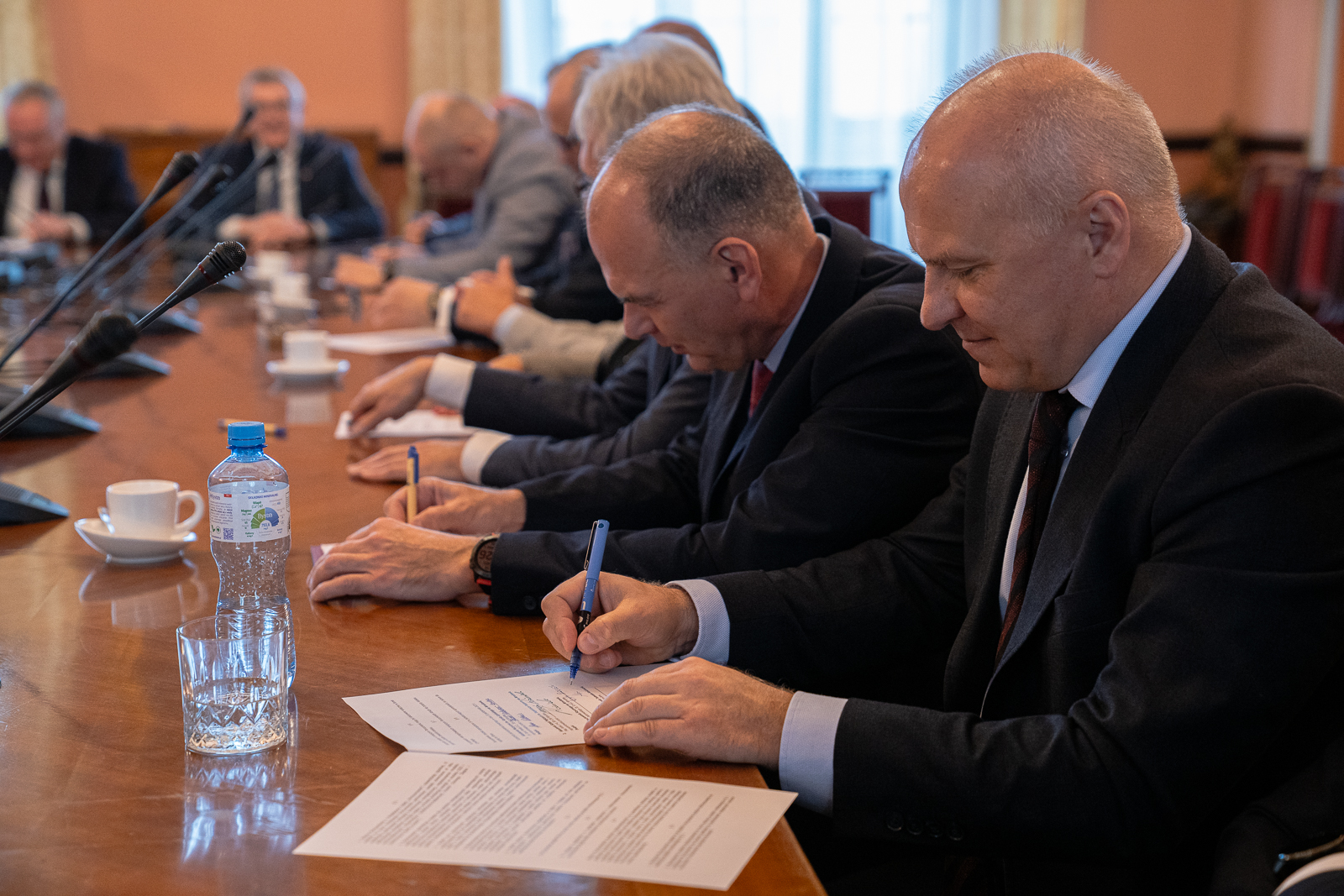 grupa mężczyzn w starszym wieku siedząca przy dużym stole , na którym stoją mikrofony, dwaj mężczyźni z przodu podpisują dokumenty