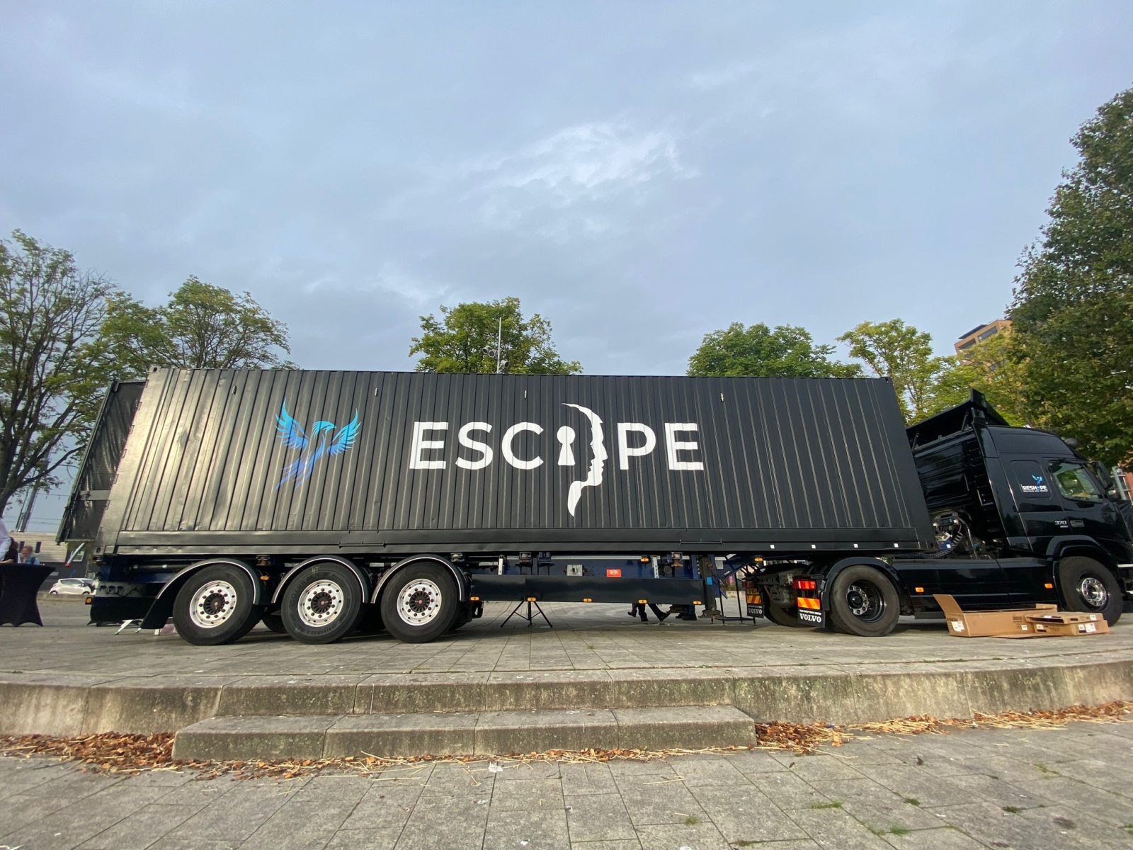 Czarny ciągnik siodłowy z czarną naczepą i kontenerem z napisem "Escape". Za nim widoczne są drzewa i wierzchołki drzew z zielonymi liśćmi.