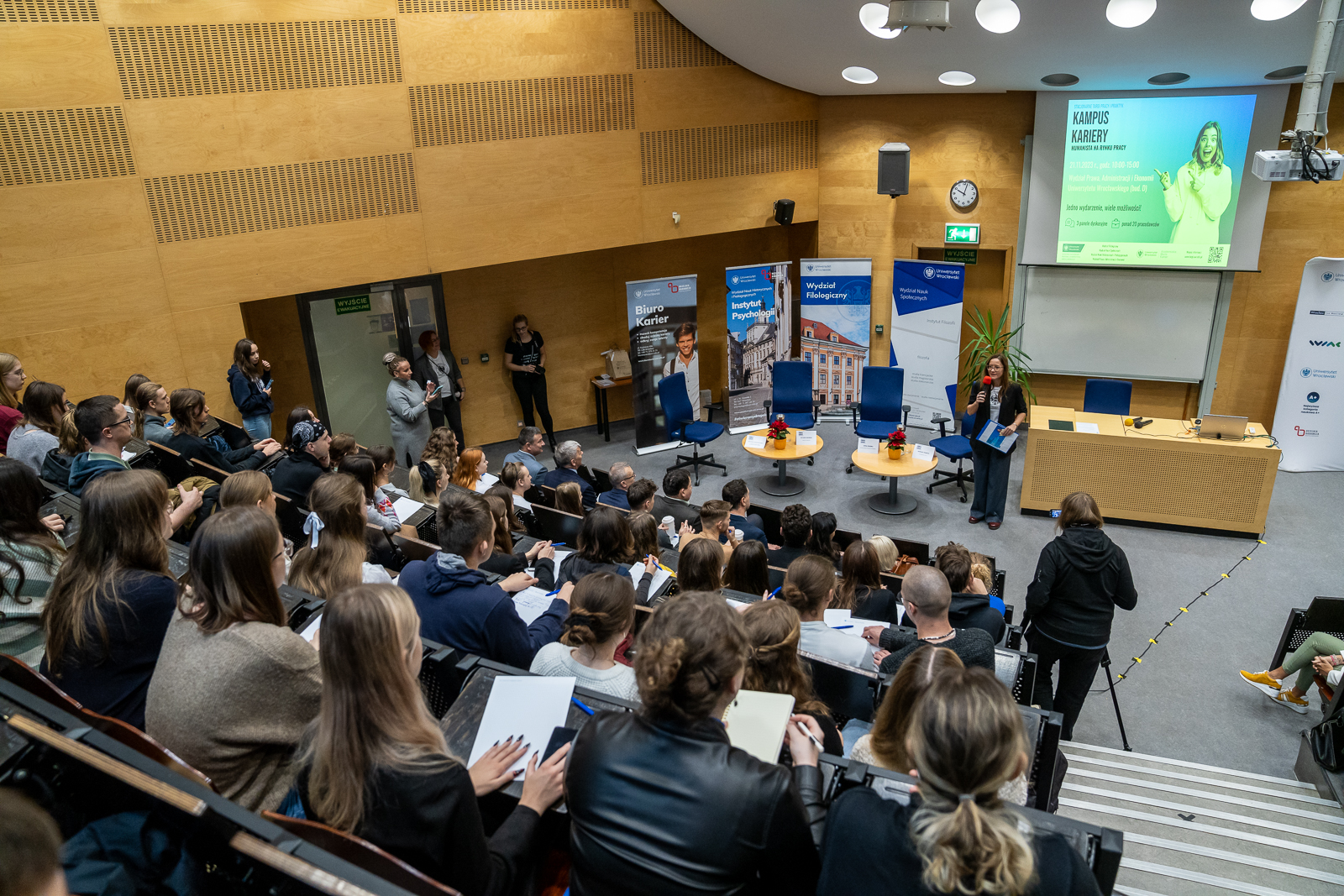 sala konferencyjna w której siedzi duża ilość osób, na środku ekran z wyświetlającymi się slajdami, pod nim młoda kobieta a mikrofonem, z lewej strony trzy niebieskie krzesła i banery reklamowe