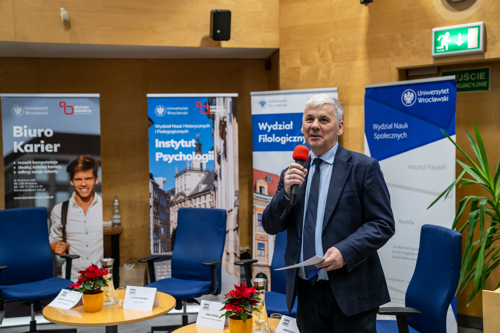 mężczyzna z mikrofonem , za nim banery reklamowe z napisem "Uniwersytet Wrocławski"