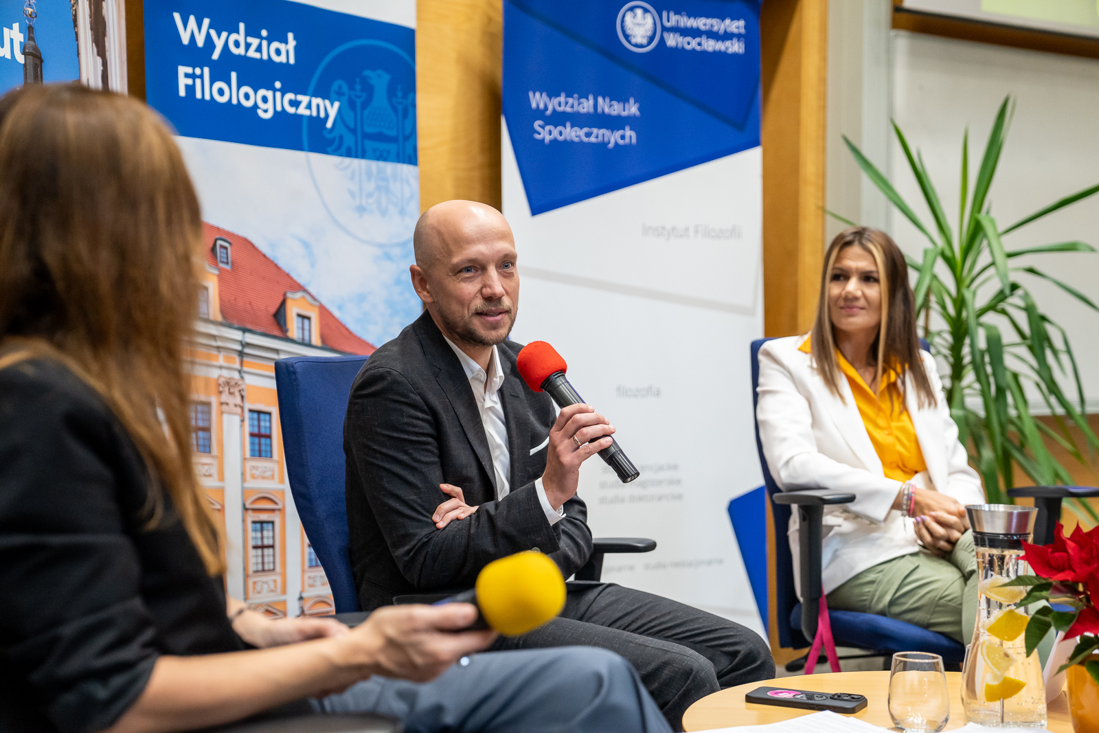 trzy osoby siedzące na krzesłach, w środku mężczyzna w średnim wieku w marynarce i białej koszuli trzymający mikrofon, z tyłu banery reklamowe z napisami "Uniwersytet Wrocławski"