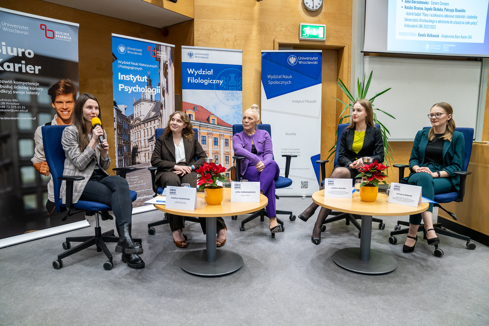 grupa pięciu kobieta, w środku starsza, wokół młode kobiety, siedzące na krzesłach, za nimi banery reklamowe, kobieta z lewej strony trzyma żółty mikrofon