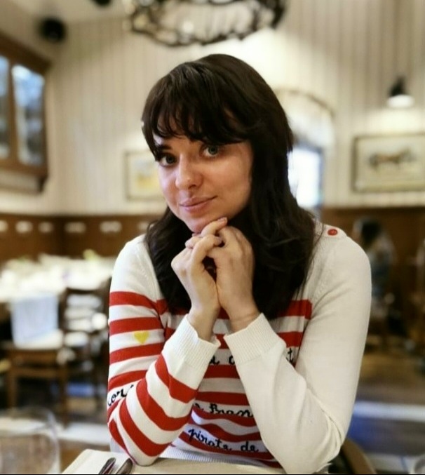 młoda kobieta o ciemnych włosach do ramion, siedzi z rękoma splecionymi pod brodą, ubrana w swetr w biało-czerwone pasy, w tle wnętrze restauracji