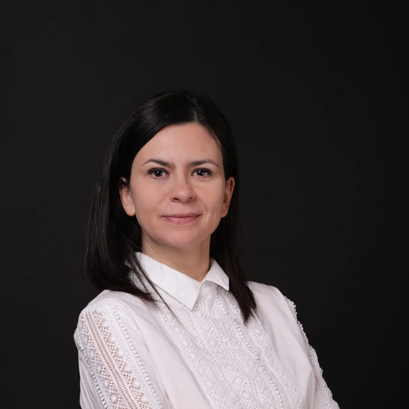 młoda kobieta o długich czarnych włosach, w białęj koszuli, stoi z założonymi rękoma