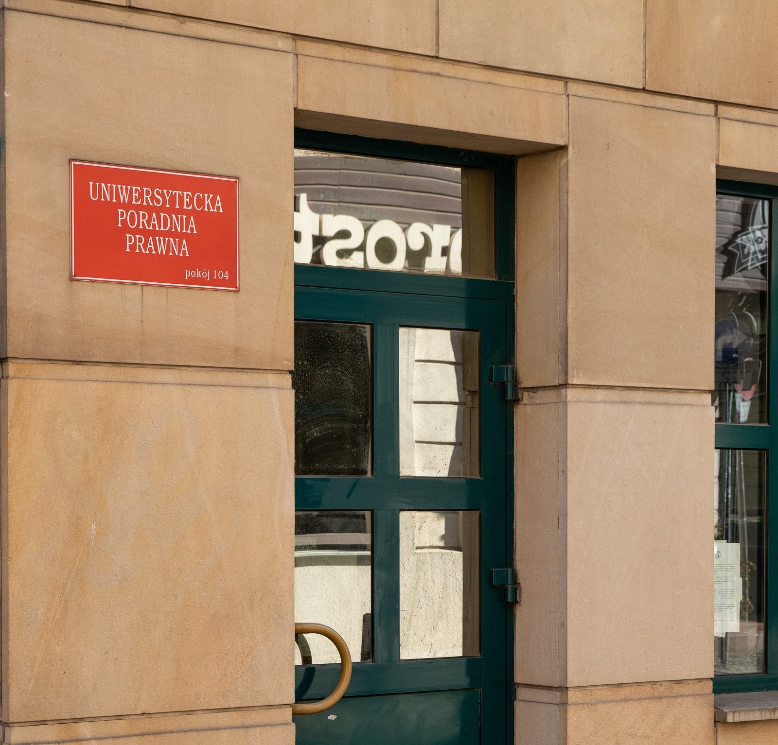 ściana budynku z czerwoną tabliczką z napisem biała czcionką "UNIWERSYTECKA PORADNIA PRAWNA" oraz z zielonymi, przeszklonymi drzwiami