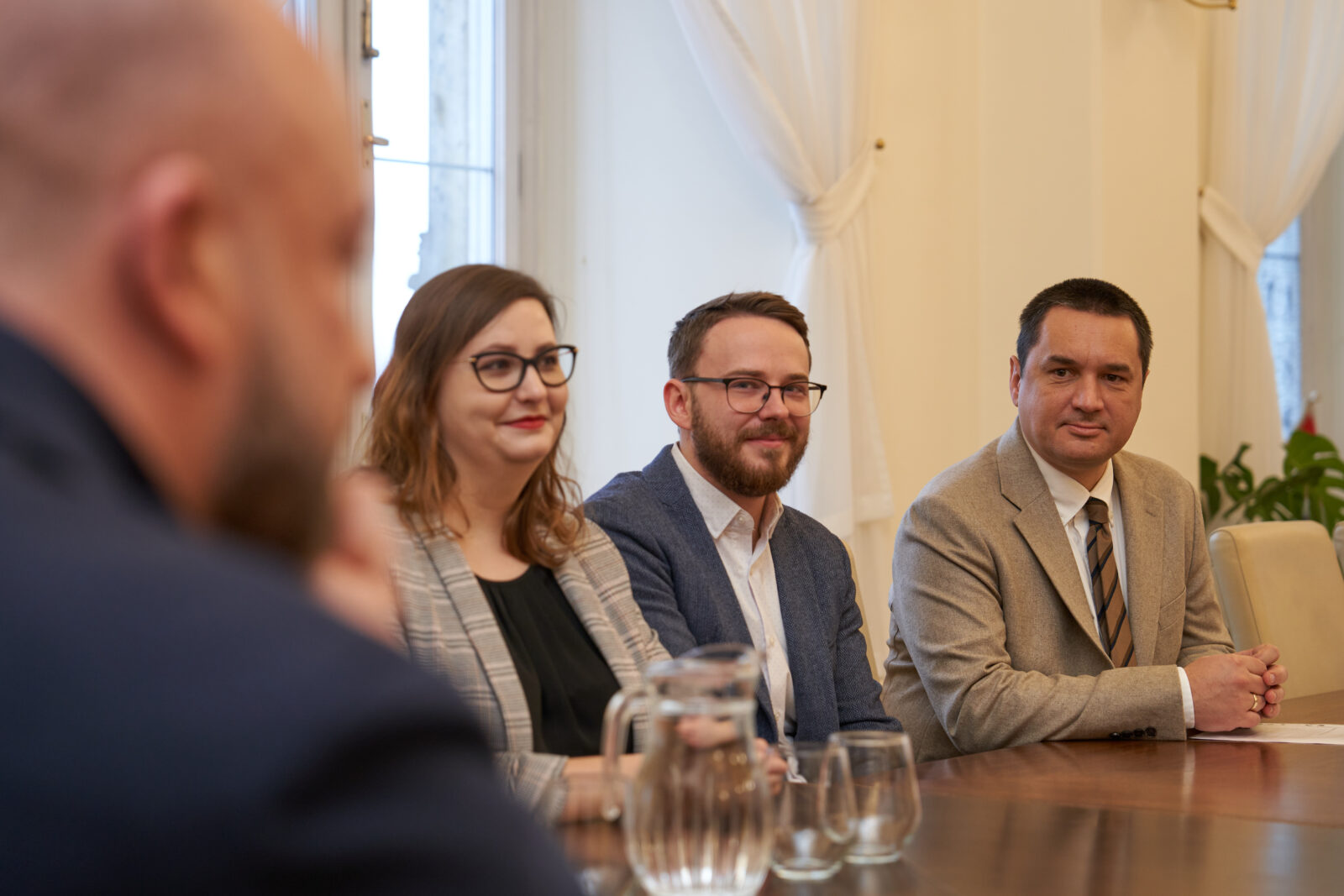 kobieta i dwóch mężczyzn przy stole, wszyscy ubrani w marynarki, z lewej strony częściowo widoczny z profilu mężczyzna, widać jego głowę i bark.