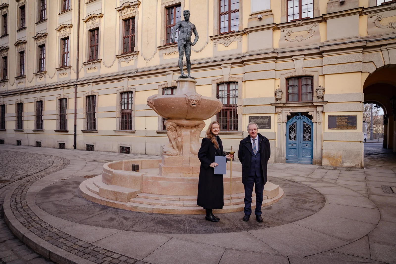 Kobieta i mężczyzna stoją przed barokowym budynkiem. Oboje ubrani są elegancko on ma na sobie garnitur, ona długi płaszcz do ziemi. Kobieta w wieku około 30 lat, ma długie włosy i jest uśmiechnięta. Trzyma w jednej ręce teczkę, a w drugiej szpadę. mężczyzna ma ręce splecione za plecami. Jego twarz jest uśmiechnięta, nosi okulary, ma krótki siwe włosy.