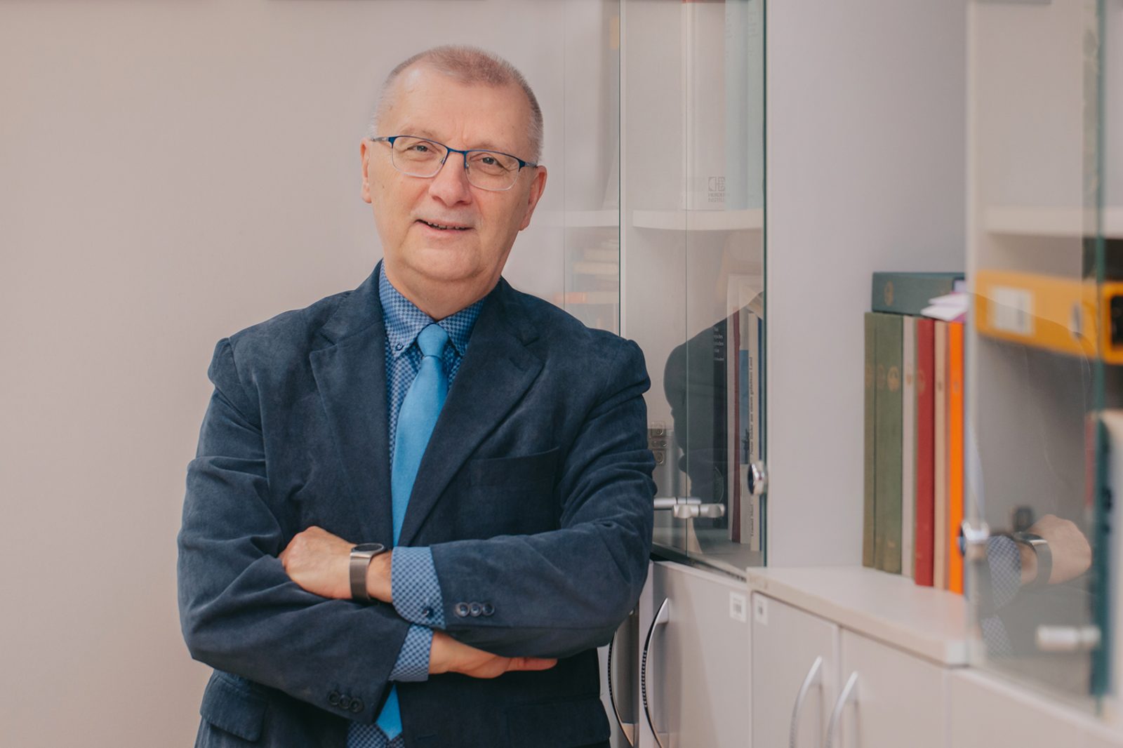 mężczyzna w średnim wieku w siwych włosach, okularach , ubrany w ciemną marynarkę, niebieską koszulę, stoi oparty o regał z książkami